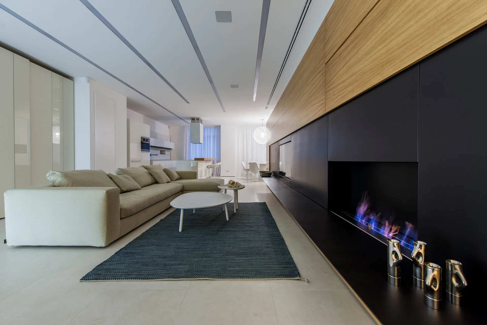 A modern living room features a beige modular sofa, a dark blue rug, and a long black media wall with an integrated fireplace.