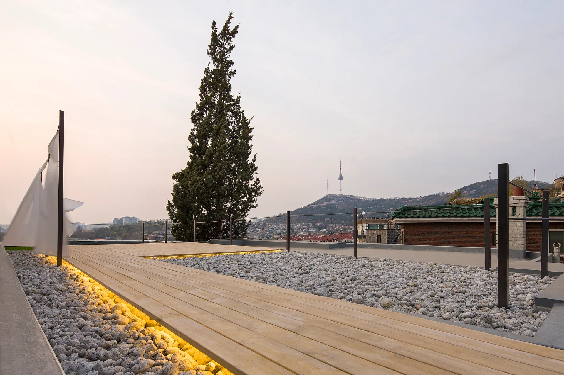 A rooftop terrace features a raised wooden plank deck with warm under-deck LED lighting, surrounded by a bed of grey gravel under a twilight sky.