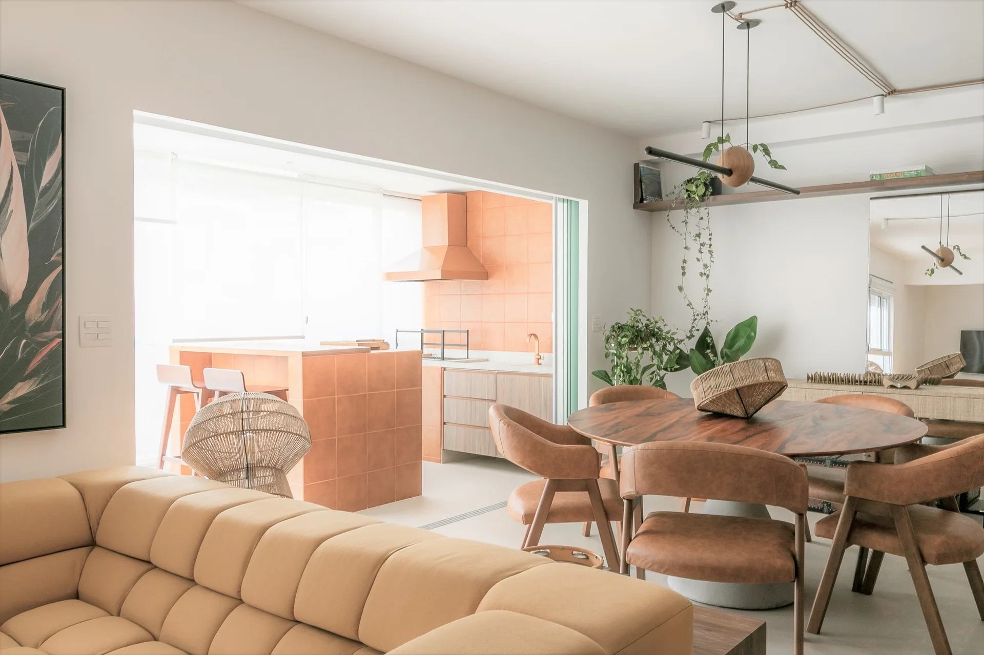 An open-concept living space features a tufted tan sofa, a round wooden dining table, and a kitchen with a terracotta-tiled island.