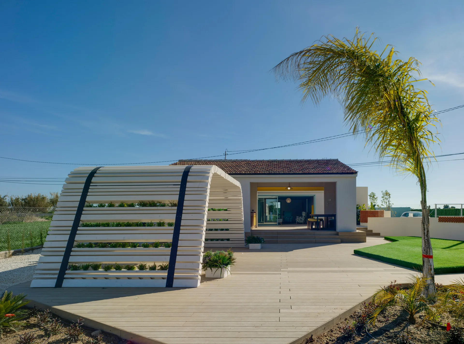 A white arched slatted pergola stands on a light wood deck next to a modern house and a tall palm tree under a clear blue sky.