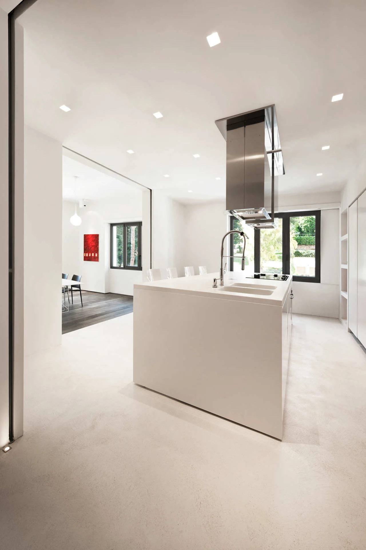 A minimalist all-white kitchen features a large central island with an integrated sink and gooseneck faucet beneath a stainless steel range hood.