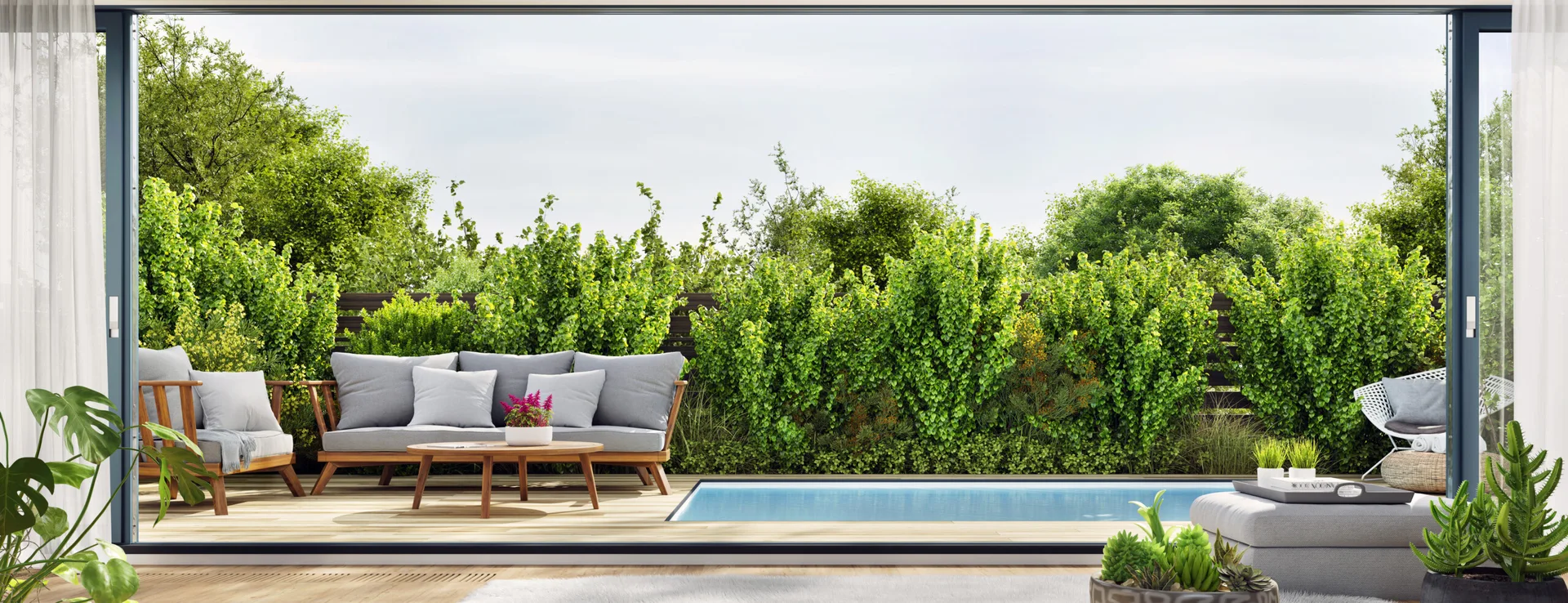 A modern wood pool deck features a grey outdoor sofa, a low wooden coffee table, and a swimming pool, all set against a tall, lush green hedge backdrop.