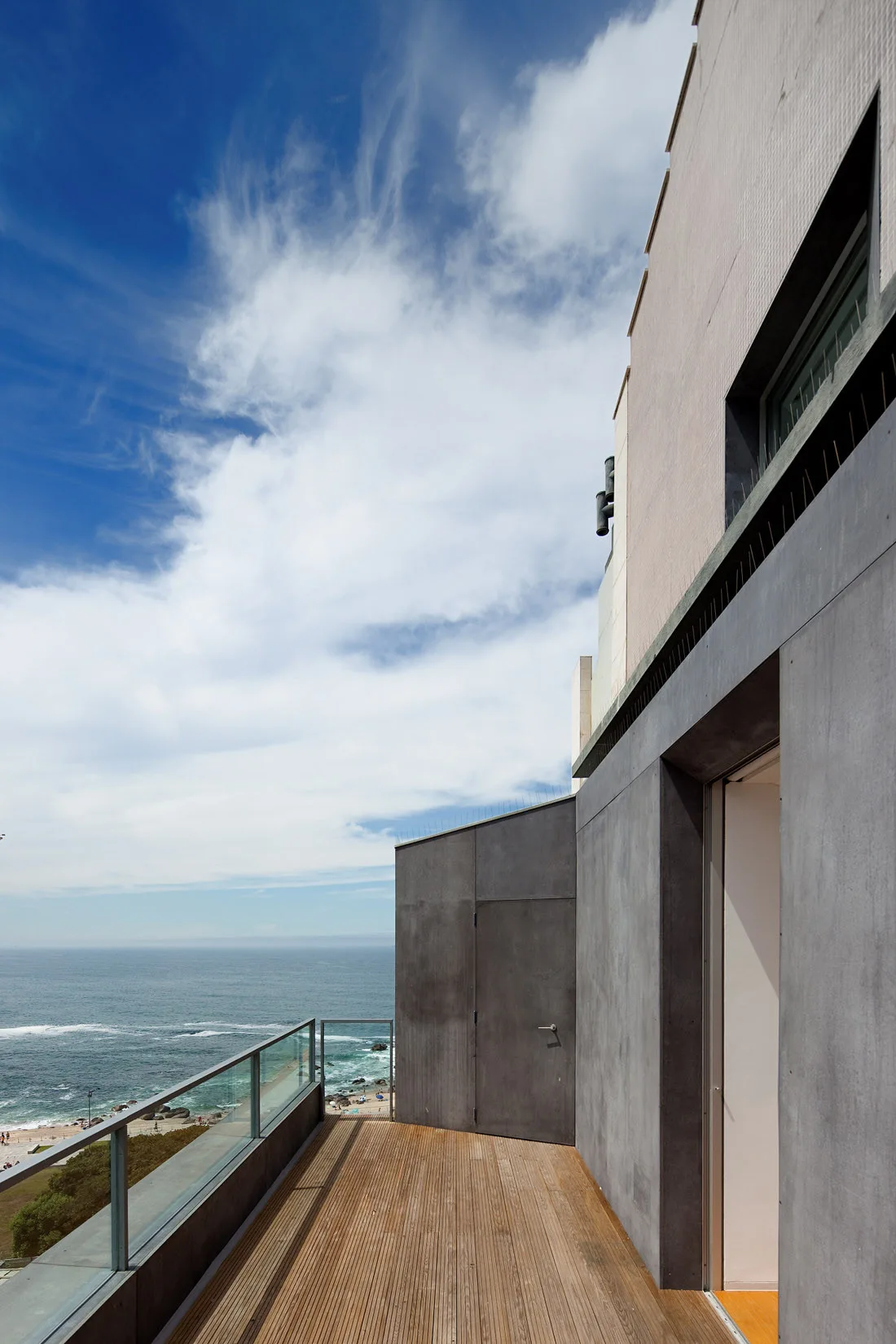 An elevated wood deck balcony with grey facade panels and a glass railing overlooking the ocean.