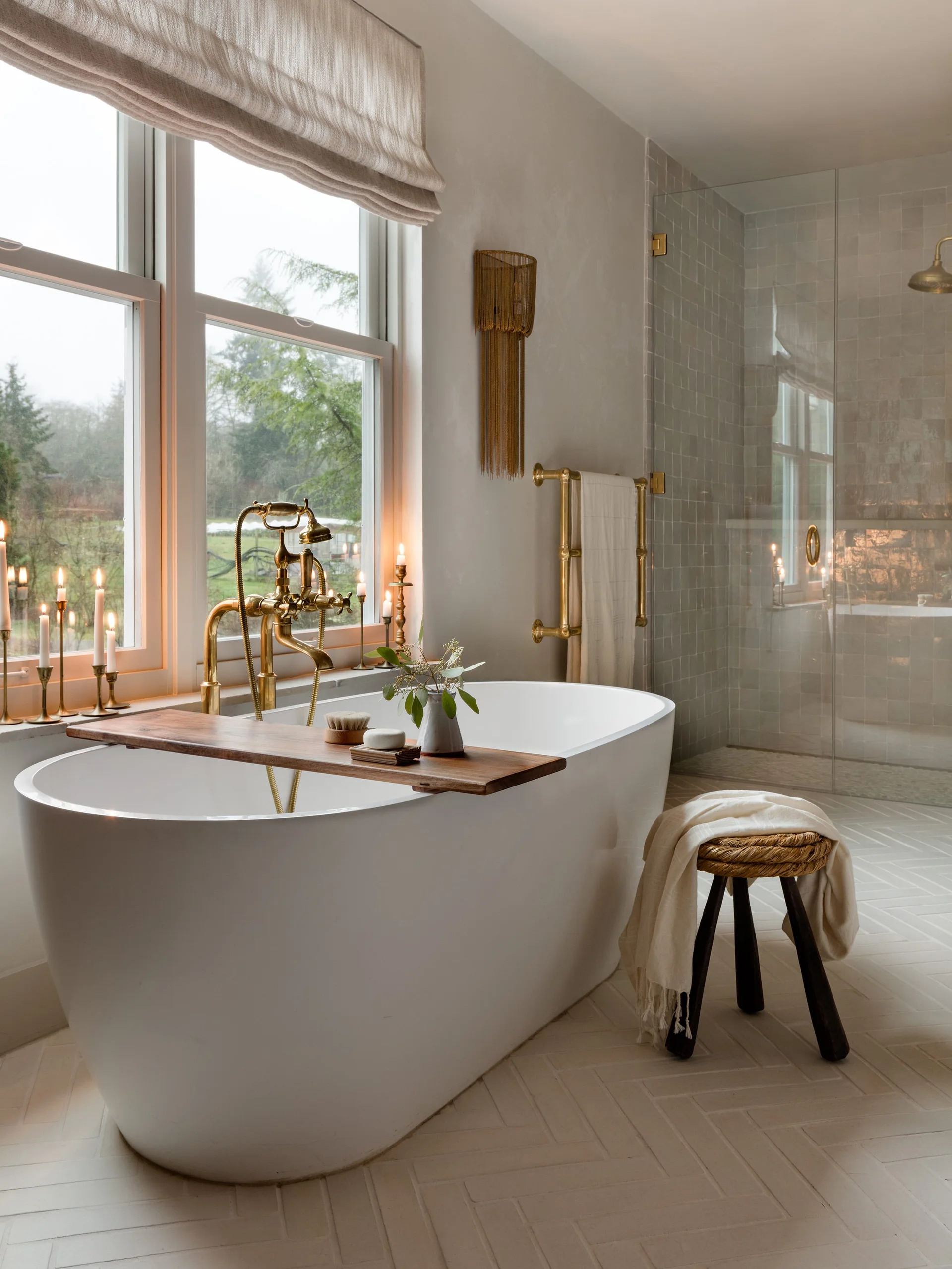 A bright bathroom features a white freestanding tub with a wooden tray, vintage brass fixtures, a small wooden stool, and a glass walk-in shower.