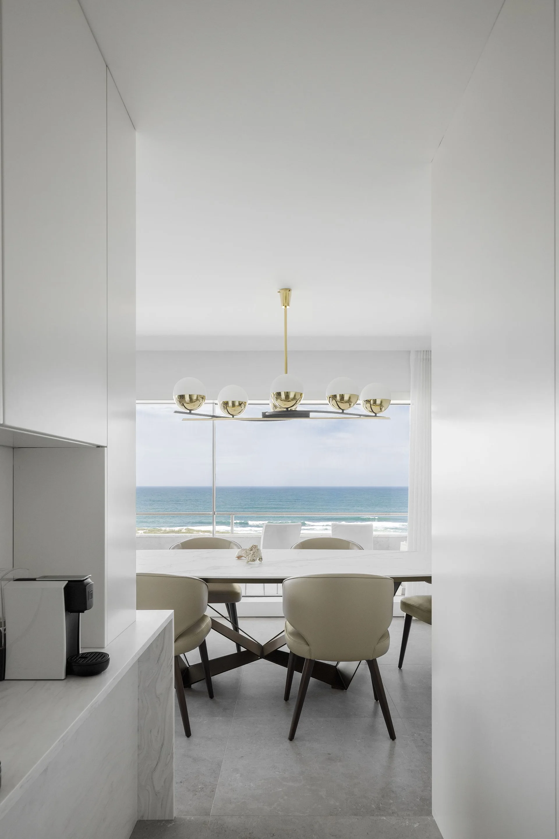 A modern dining room with a marble table, beige chairs, and a brass chandelier is framed by white cabinets and overlooks the ocean.