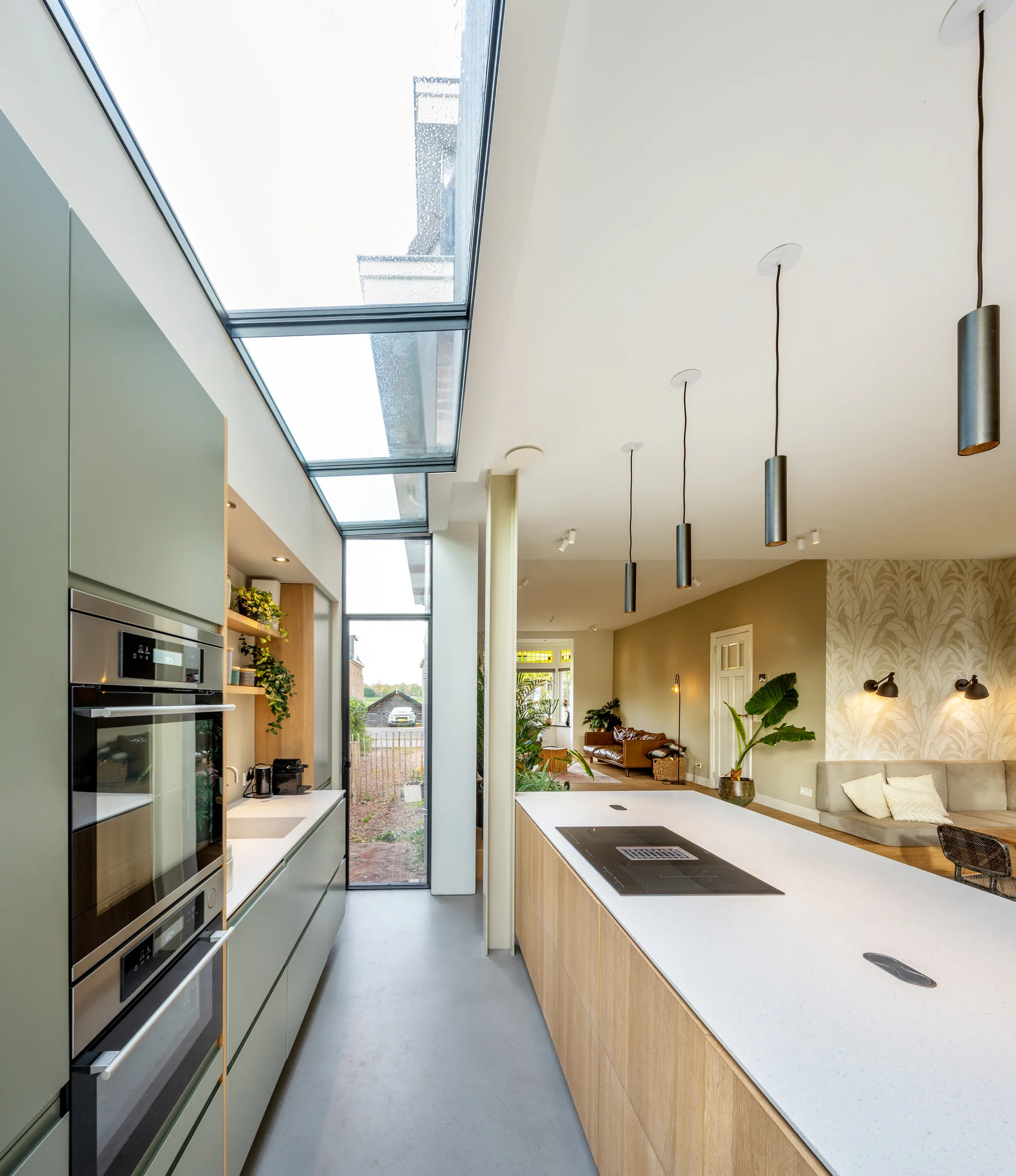 An open kitchen with sage green cabinets, a wood-front island, and a long glass skylight running above a concrete floor.