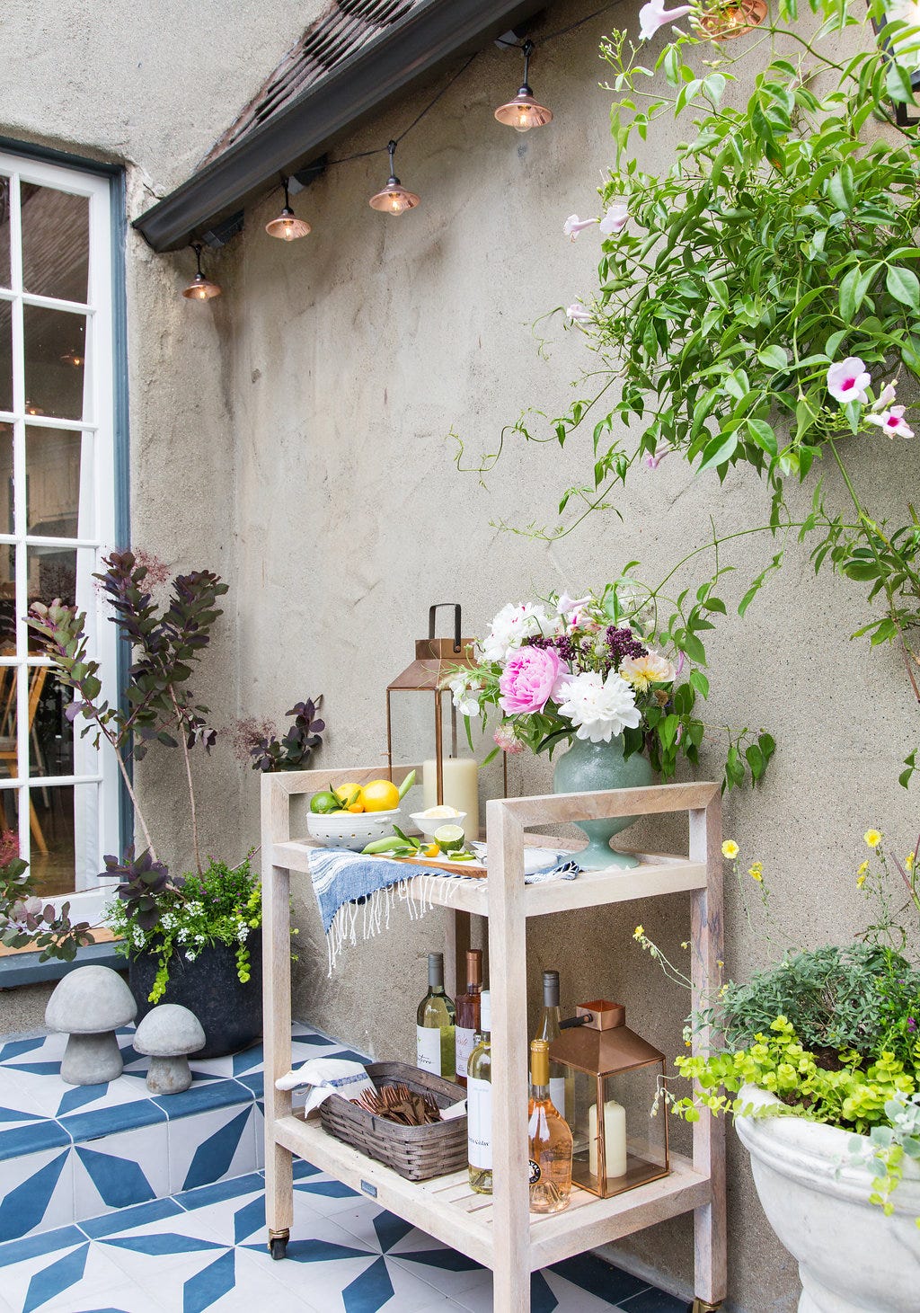 A wooden bar cart styled with flowers and wine sits on blue and white geometric patio tiles next to a textured stucco wall.