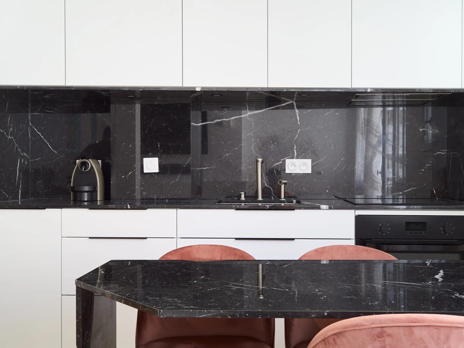 A minimalist kitchen features white flat-front cabinetry, a black veined marble backsplash, a matching dining table, and dusty pink velvet chairs.