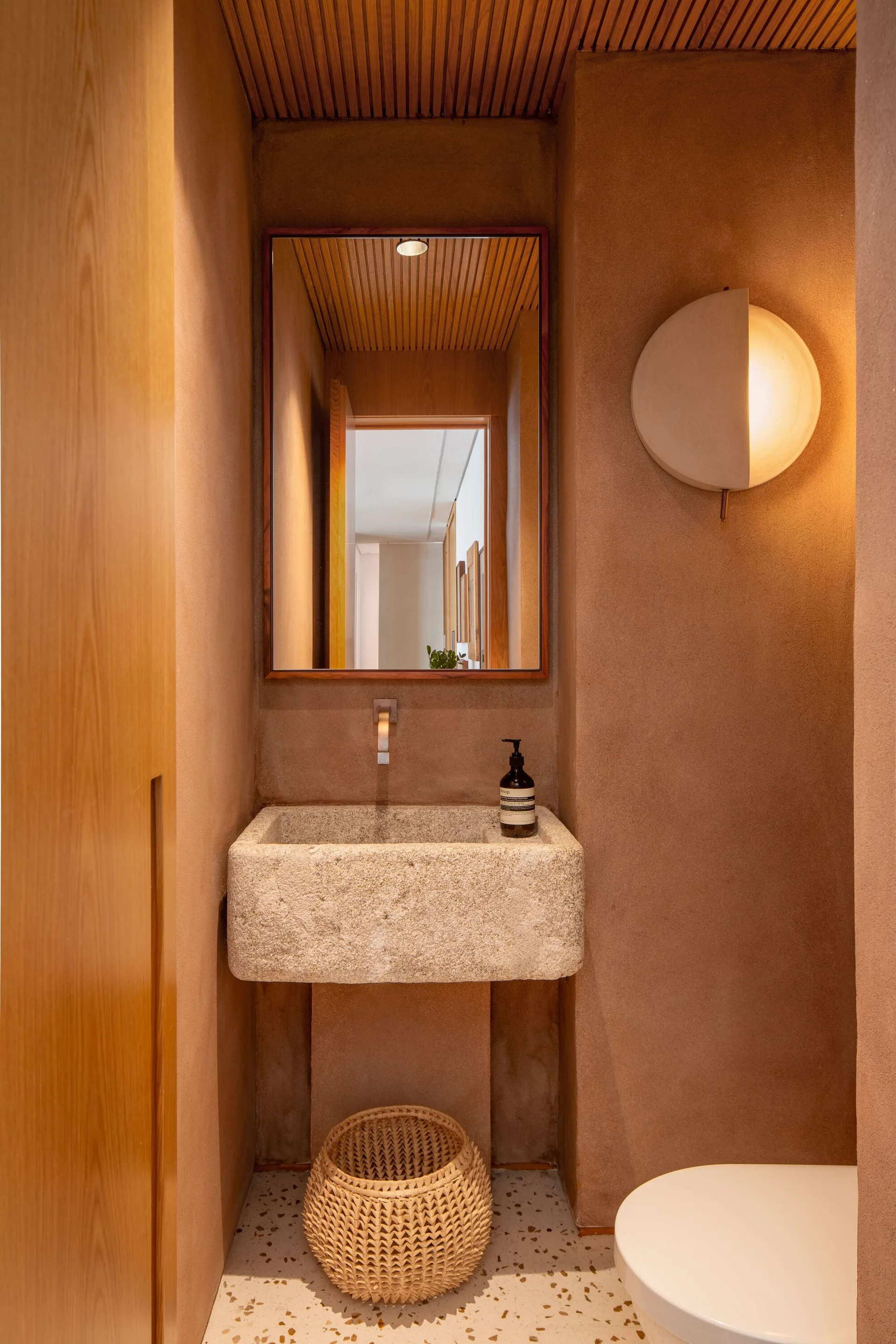 A small powder room features textured brown plaster walls, a rough stone basin sink under a rectangular mirror, and a slatted wood ceiling.