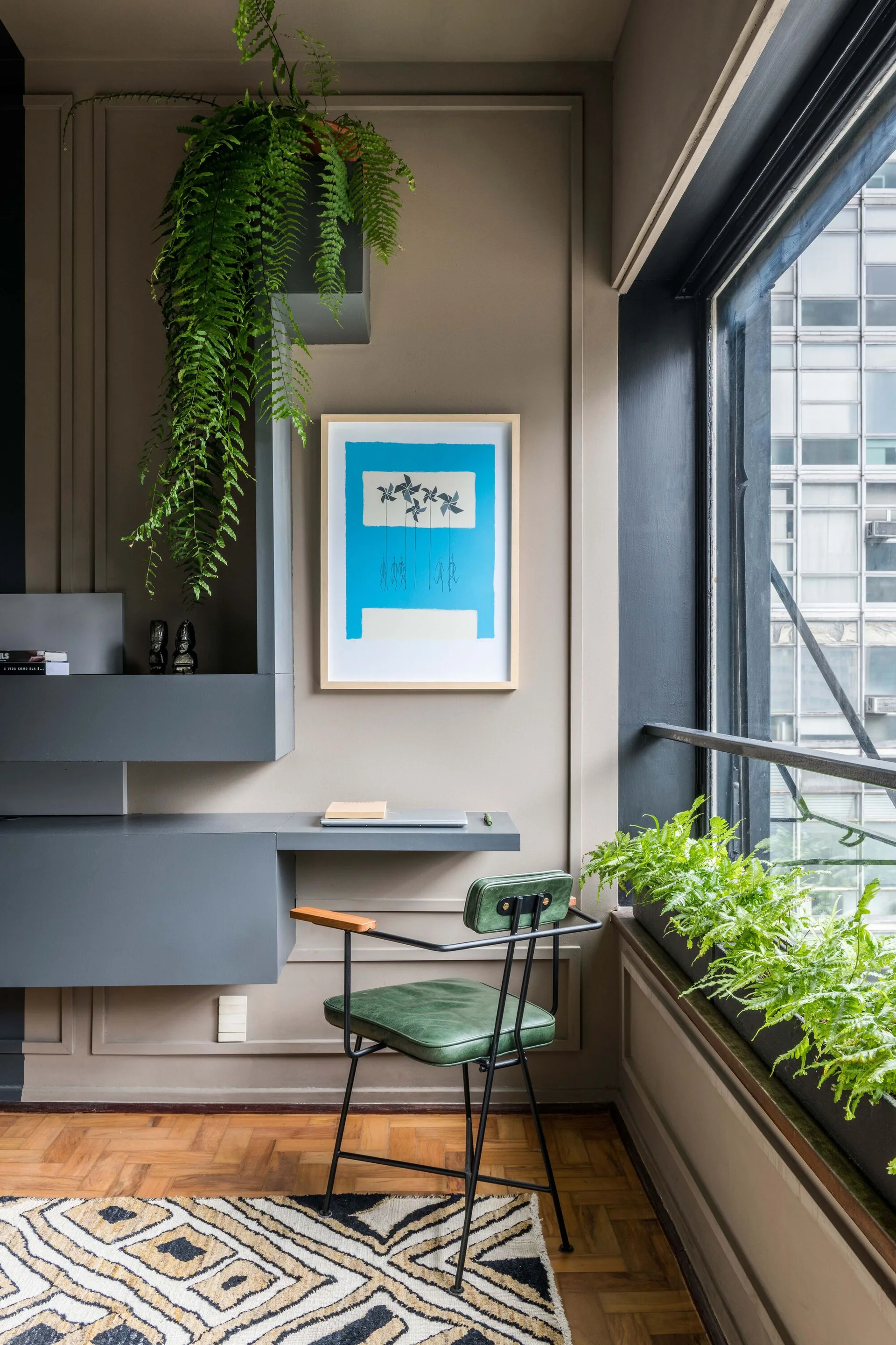 A compact home office features a floating gray desk, a green leather chair, a hanging fern, and a framed blue art piece on a taupe paneled wall.