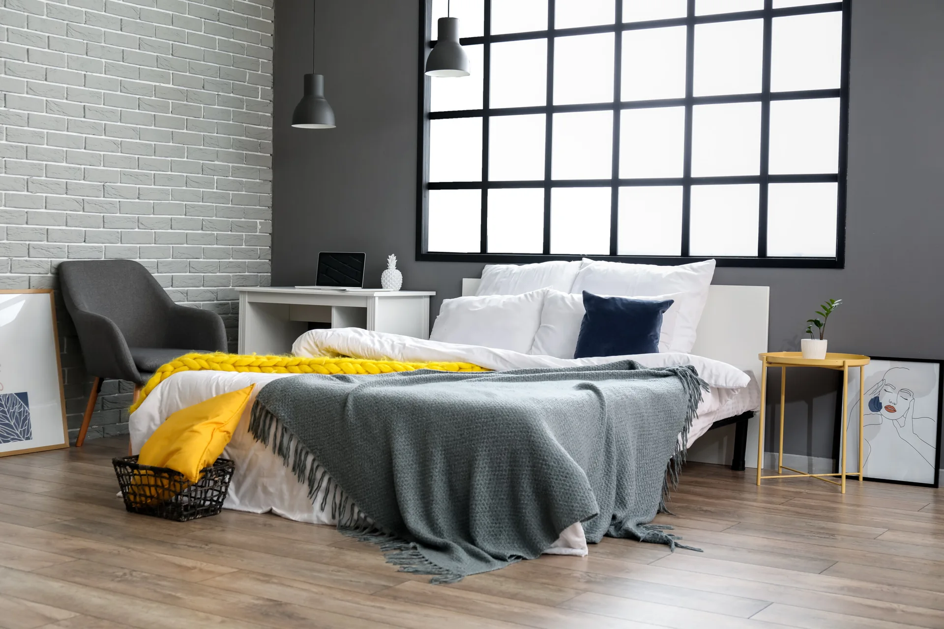 A modern bedroom features a grey brick wall, a black grid window, white bedding with yellow and grey throws, and a yellow side table.