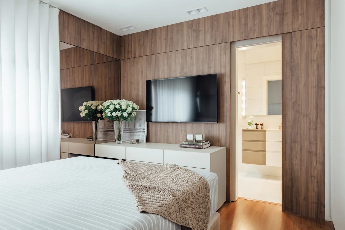 A modern bedroom features a warm wood-paneled wall, a wall-mounted TV over a low beige dresser, and an open doorway leading to a bright bathroom.