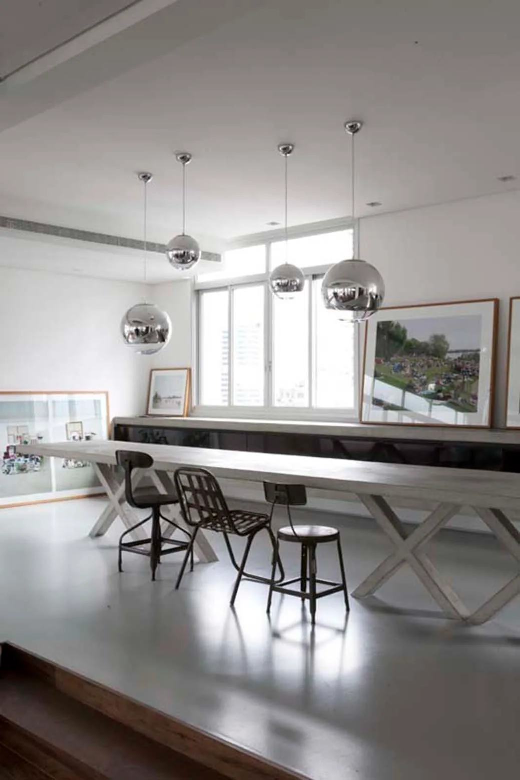 A long light-wood trestle dining table is paired with industrial stools and illuminated by shiny spherical chrome pendant lights hung in a line.