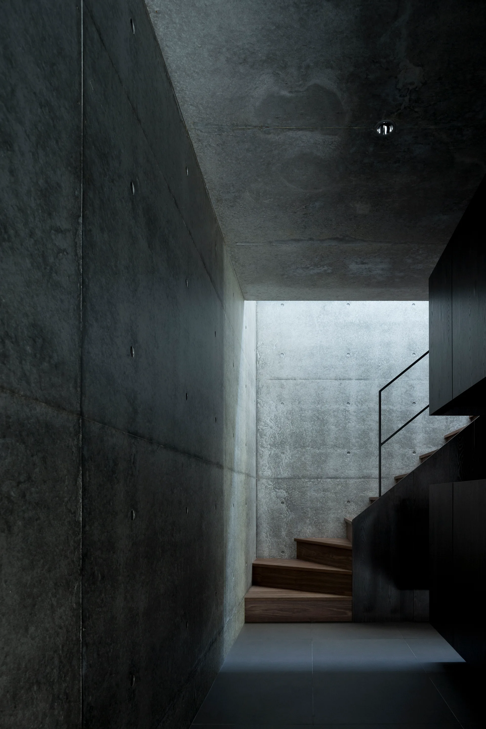 A minimalist staircase features warm wooden treads, raw concrete walls, a dark handrail, and a skylight casting light from above.