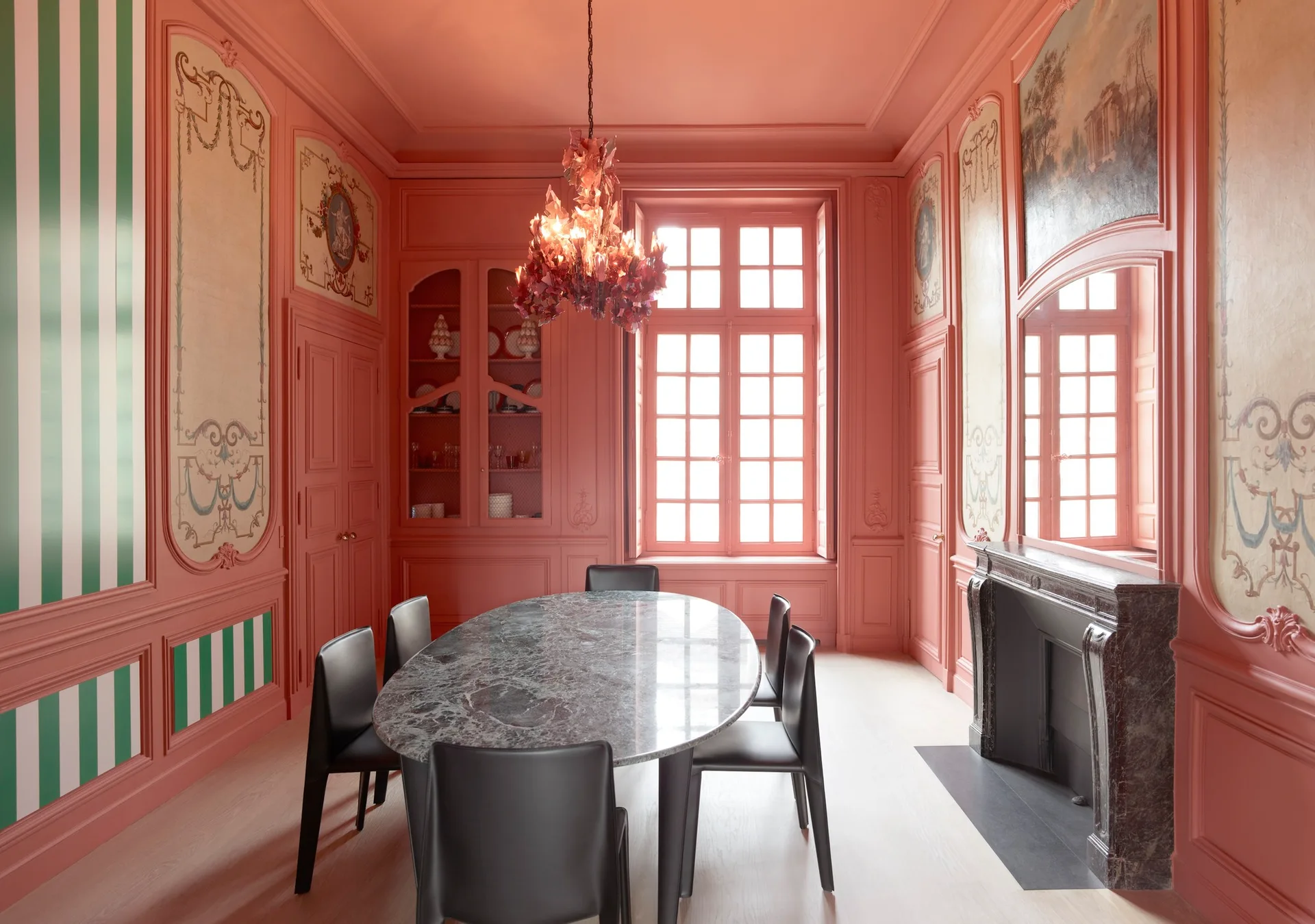 A coral-pink dining room features a marble oval table, black leather chairs, ornate wall paneling, and a red glass chandelier hanging above.