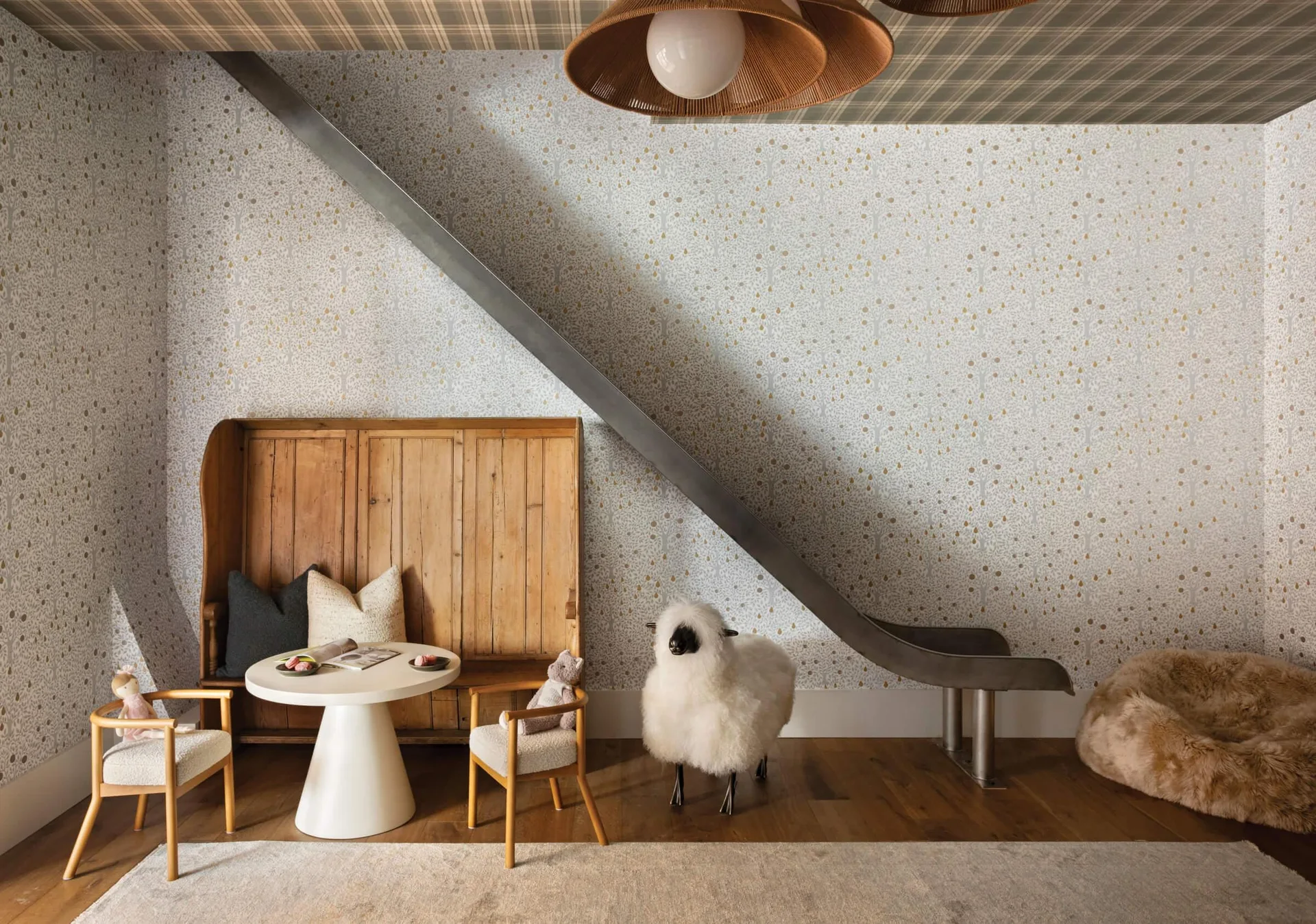 A whimsical kids' playroom featuring a diagonal metal slide, a wooden settle bench with a small round white table, tree-motif wallpaper, and a plaid ceiling.