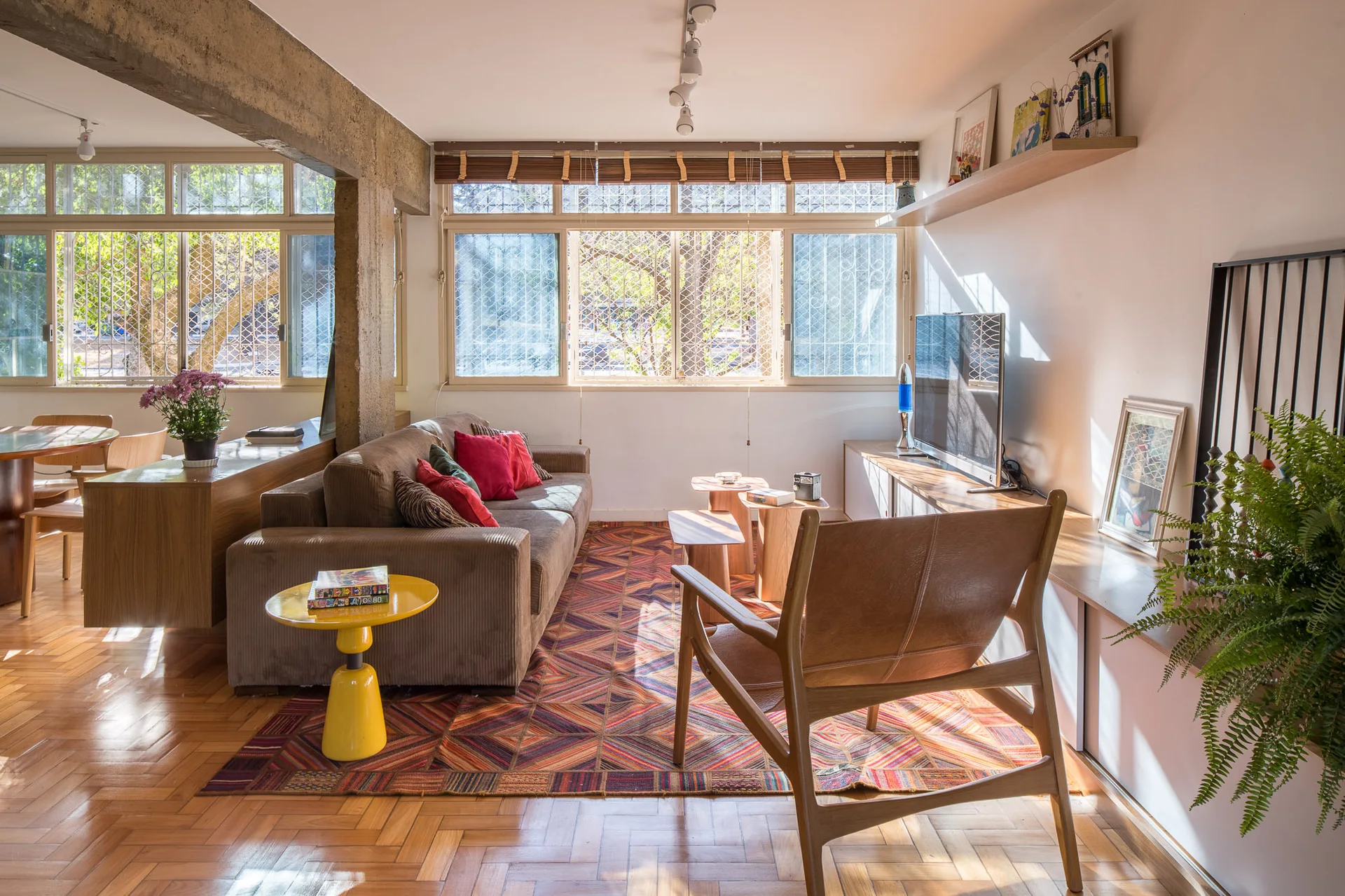 A mid-century modern living room with a brown sofa, a leather armchair, a geometric rug, and an exposed concrete beam overhead.