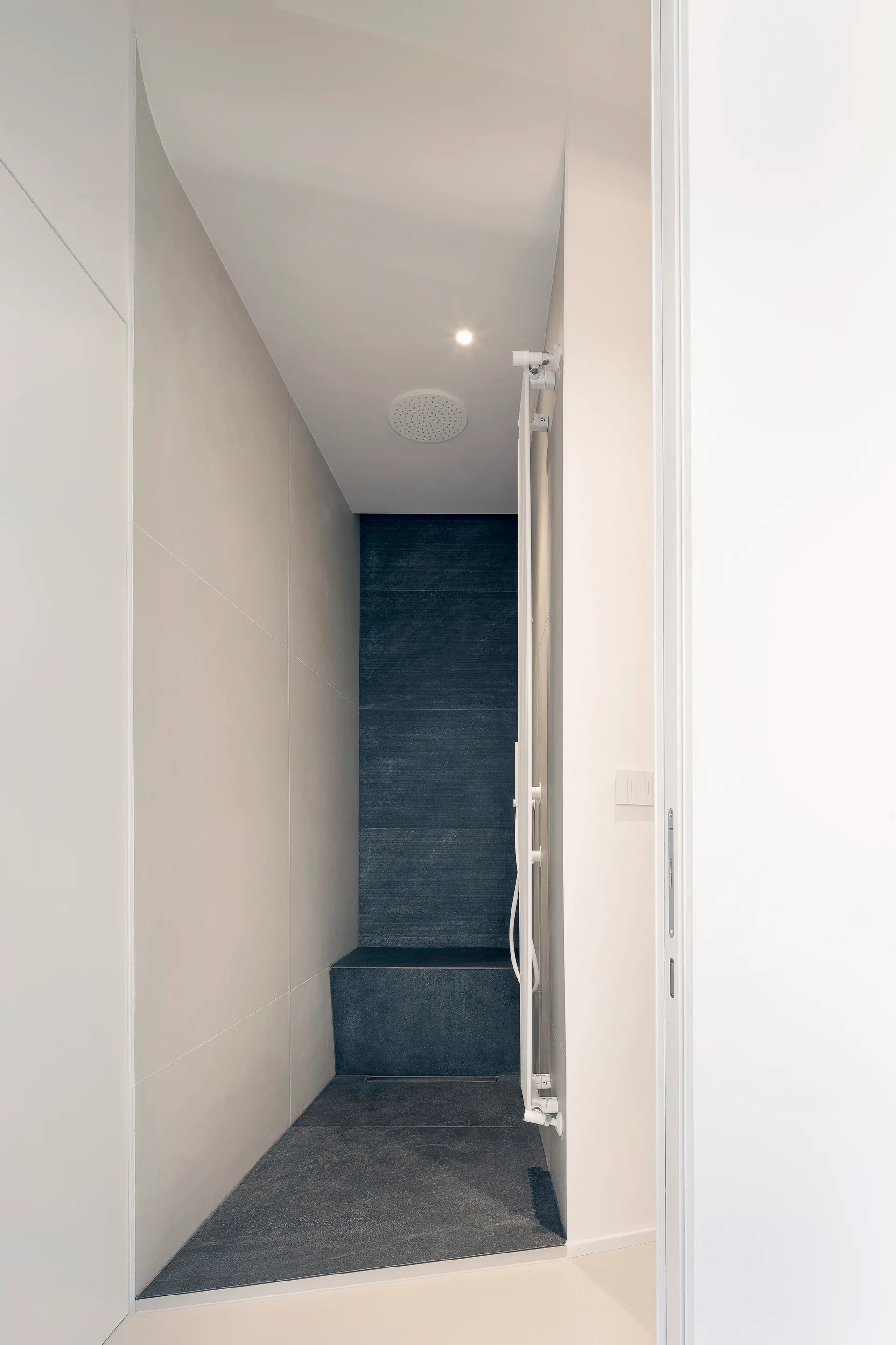 A narrow walk-in shower featuring large beige wall tiles, a dark charcoal tiled bench and floor, and a white vertical towel warmer.