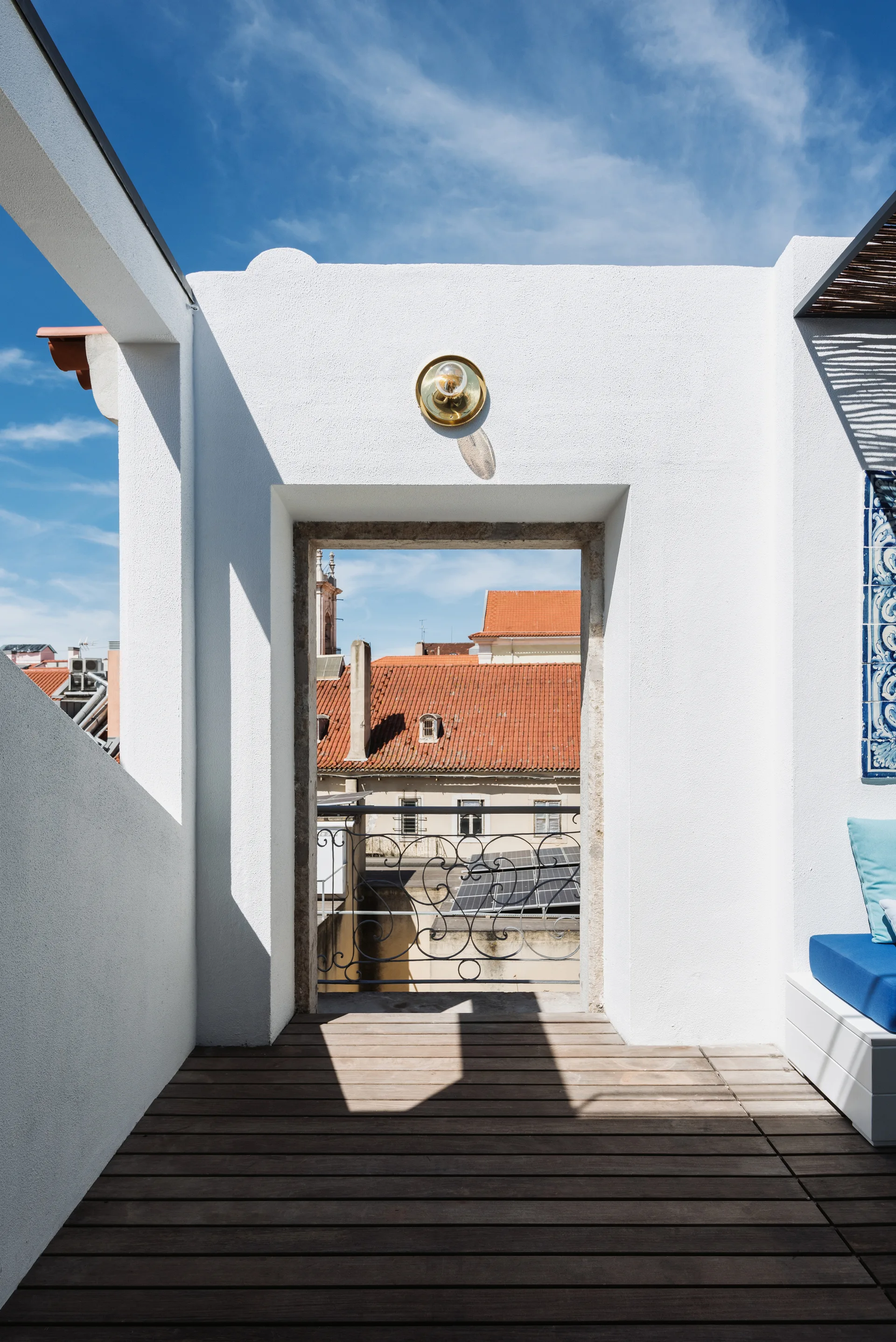 A white stucco terrace with wooden plank flooring features an open doorway framing a view of historic tiled roofs beyond a wrought iron railing.