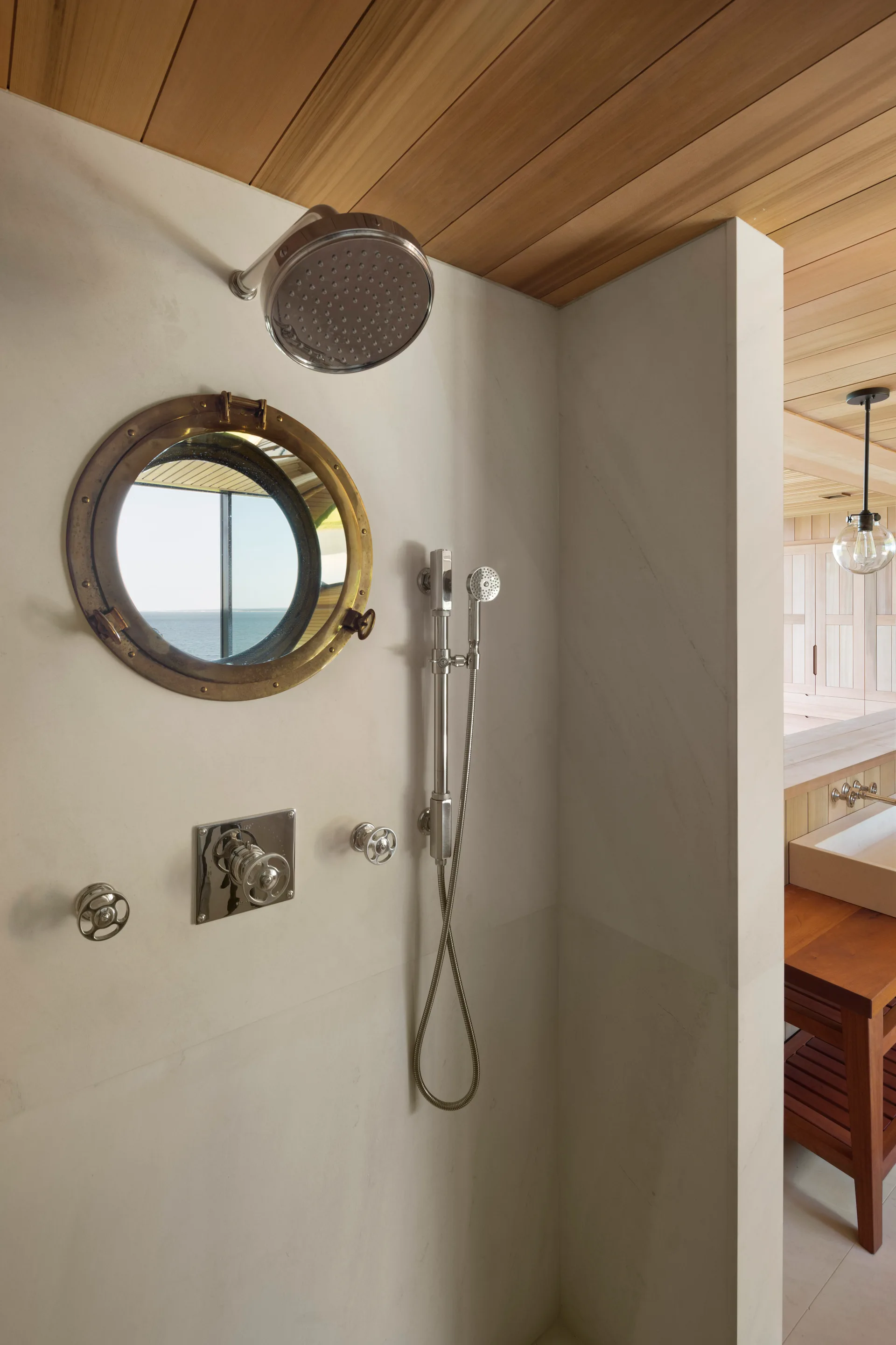A cream stone shower features a brass-framed nautical porthole window, polished silver fixtures, and a warm wood plank ceiling.