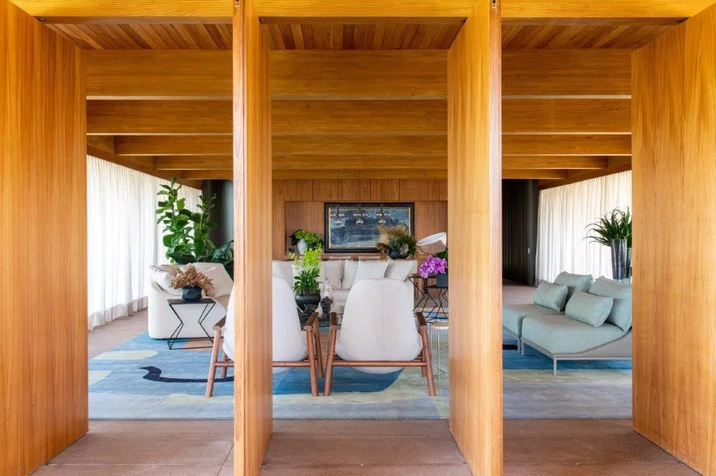 Vertical wood partition columns frame a view into a warm living room with white seating, a patterned blue rug, and sheer curtains.