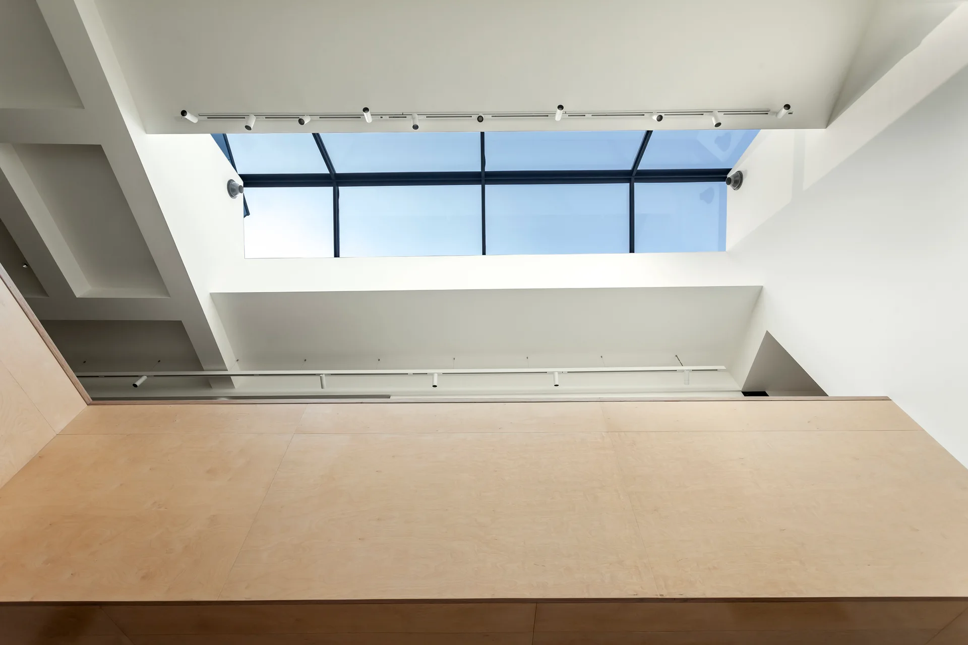 Looking up at a large rectangular skylight set into a white ceiling above light-colored plywood wall paneling with track lighting.
