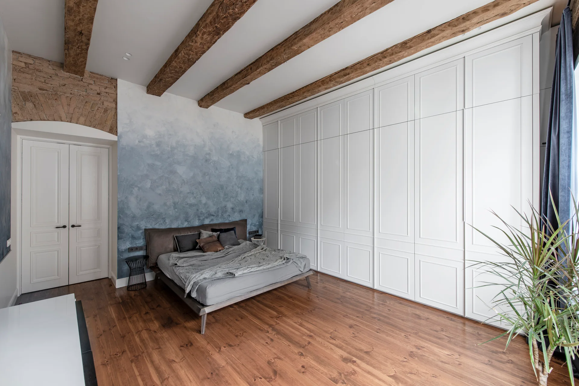 A bedroom features a blue ombre accent wall, a low grey bed, exposed wooden ceiling beams, and a large white paneled wardrobe.