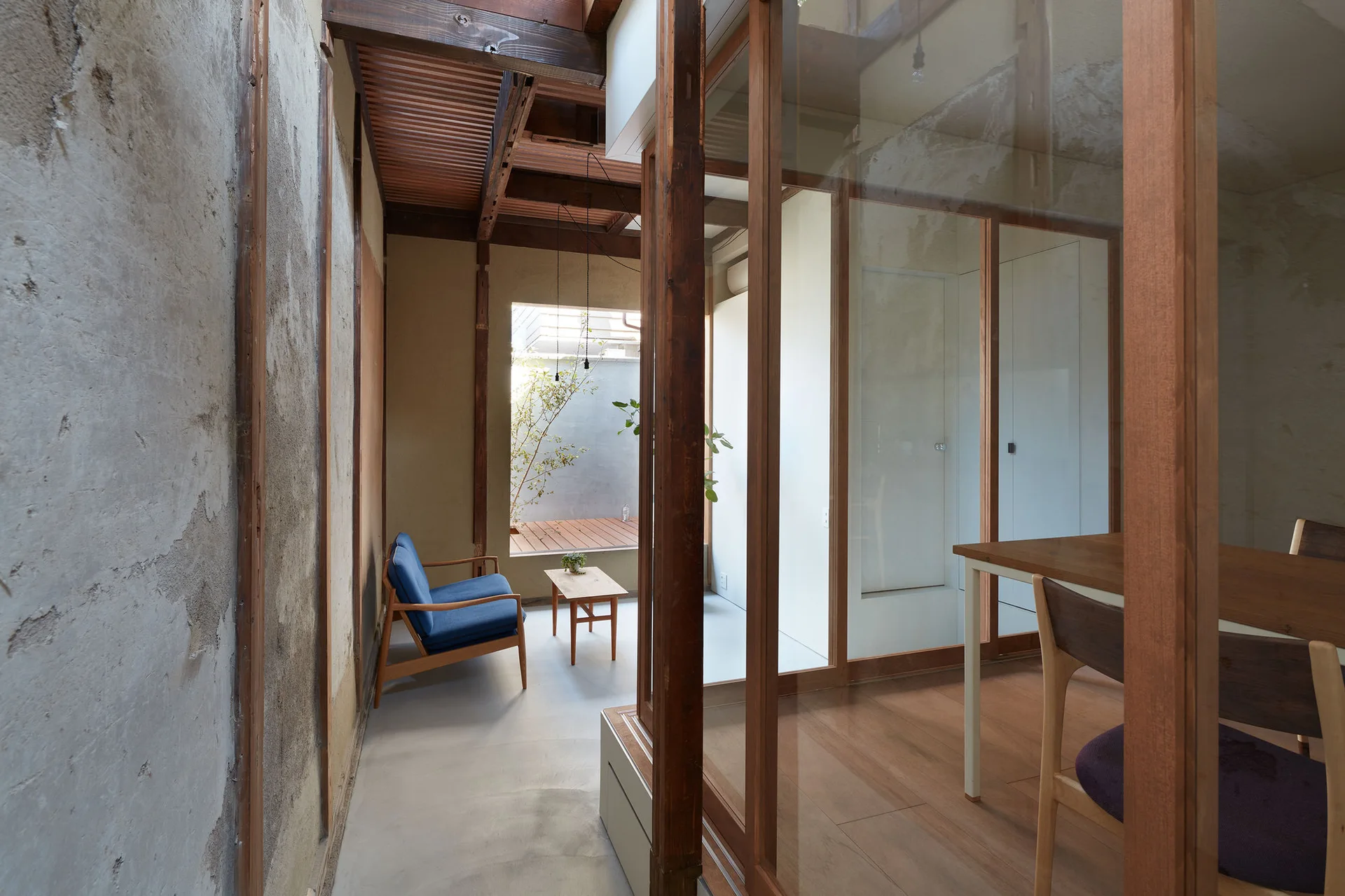 A minimalist interior featuring exposed timber beams, a blue armchair, a small side table, and glass walls looking into a dining area and courtyard.