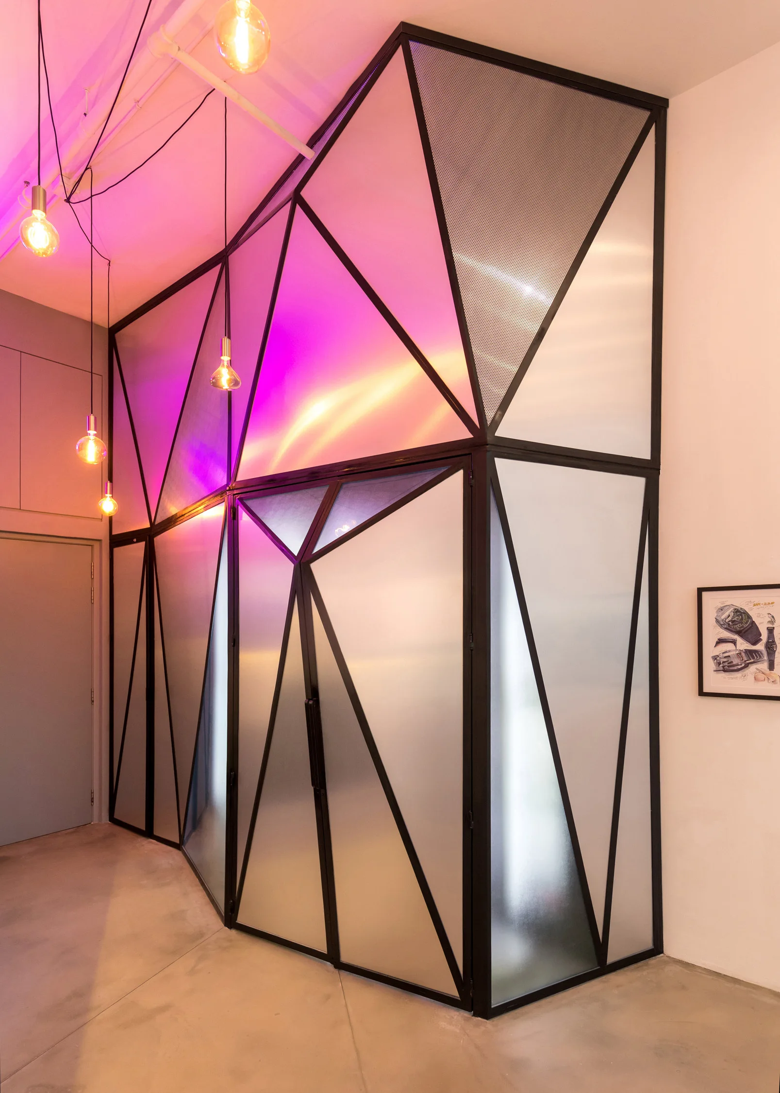 A geometric partition wall constructed of black metal and frosted glass panels stands on a concrete floor illuminated by hanging bulb lights.
