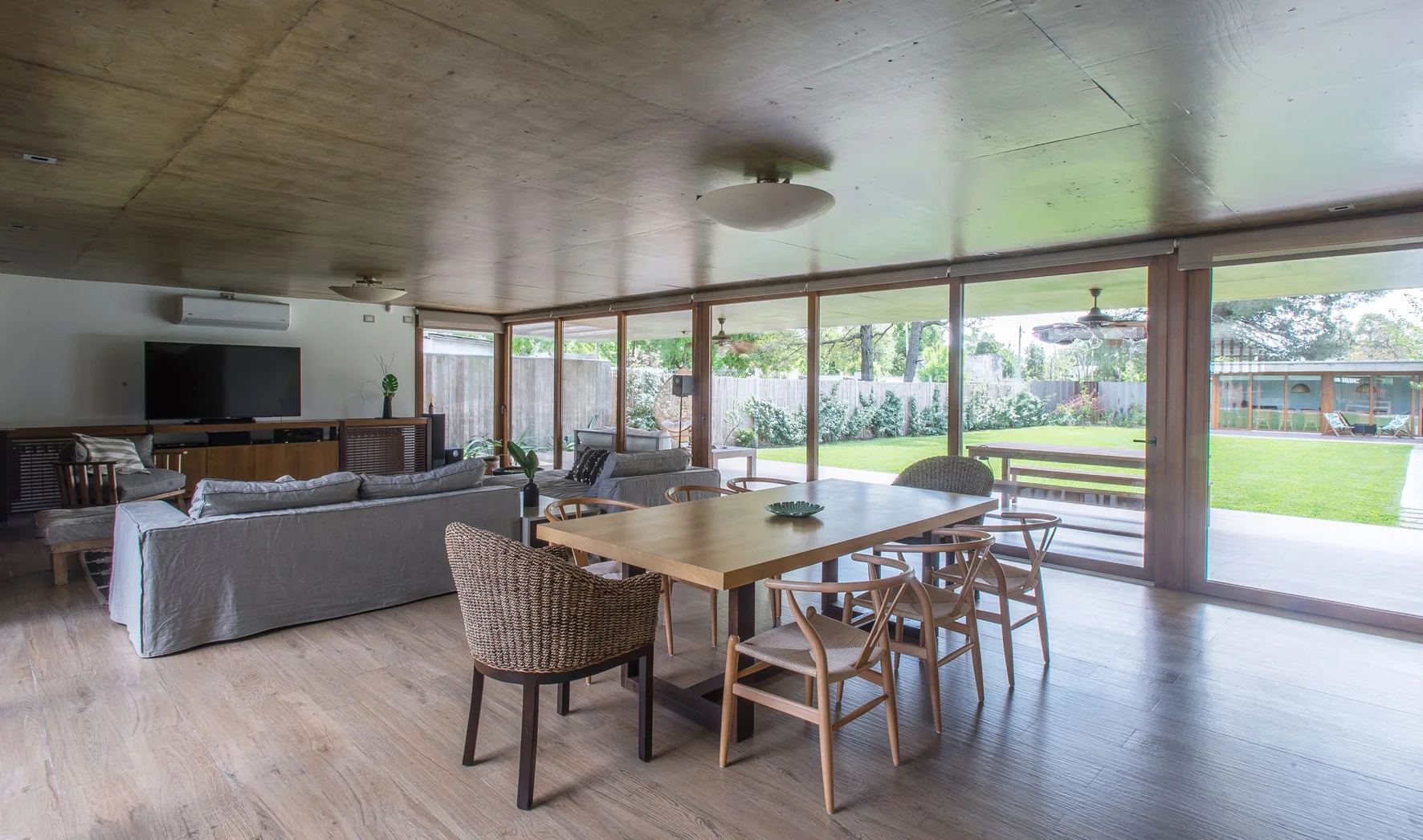A wood-paneled dining and living area features a large wooden table with chairs, a grey sofa, and sliding glass doors opening to a grassy lawn.