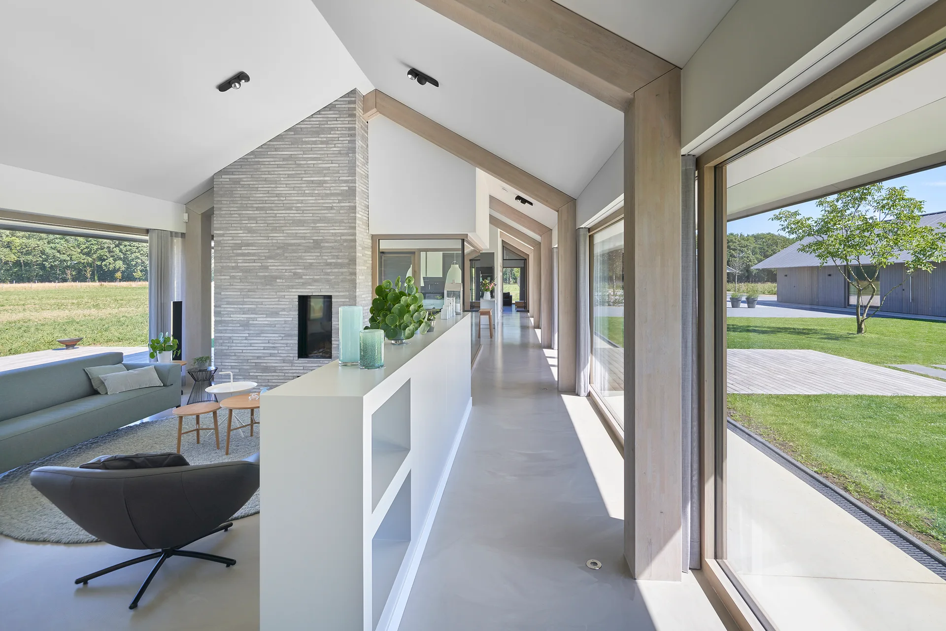 An open-plan living room featuring a vaulted ceiling with light wood beams, a low white partition divider, and floor-to-ceiling glass windows.