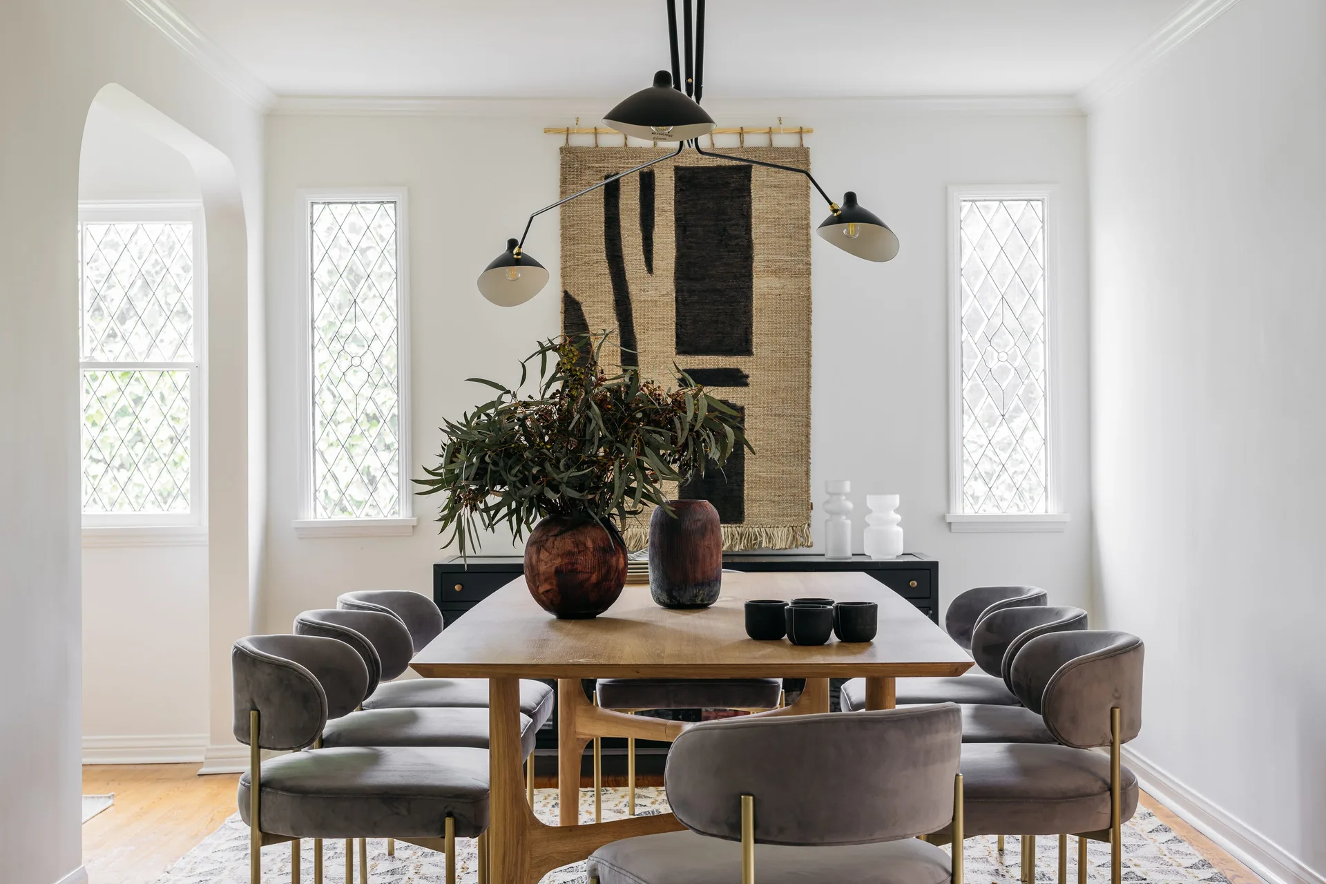 A modern dining room with a wooden table, grey velvet chairs, a three-arm black pendant light, and a large woven tapestry hanging between two lattice windows.