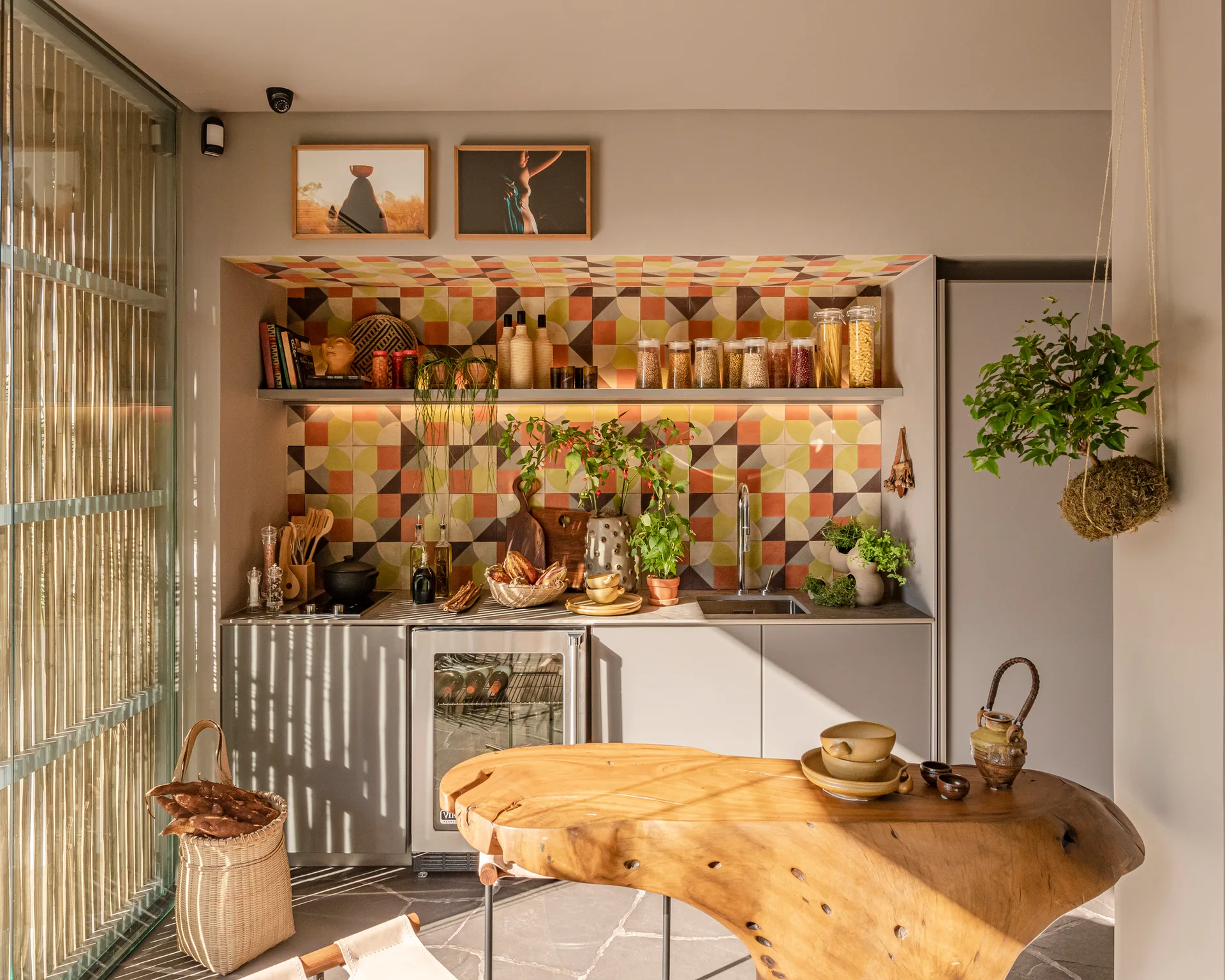A compact kitchen features a colorful geometric tile backsplash, gray cabinets, and a prominent live-edge wood island table on a stone floor.