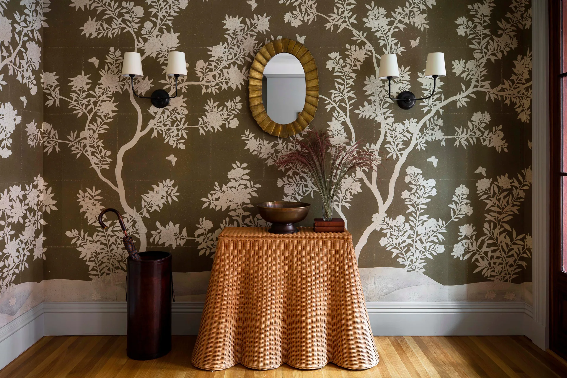 A woven wicker console table with a scalloped edge stands against olive green floral wallpaper in an entryway under two lit wall sconces.