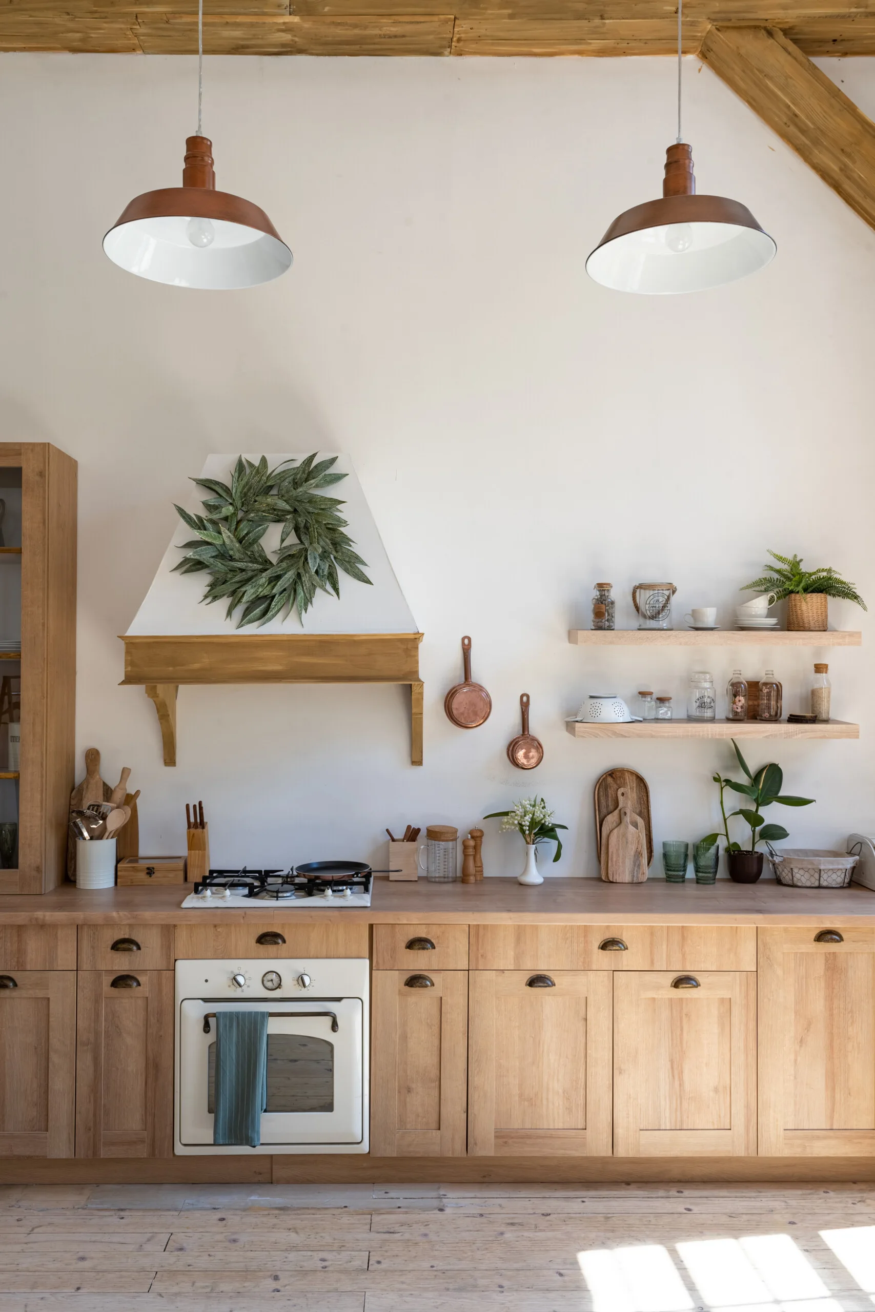 A rustic kitchen features natural wood cabinets, floating shelves with glassware, a white range hood decorated with a green wreath, and pendant lights.