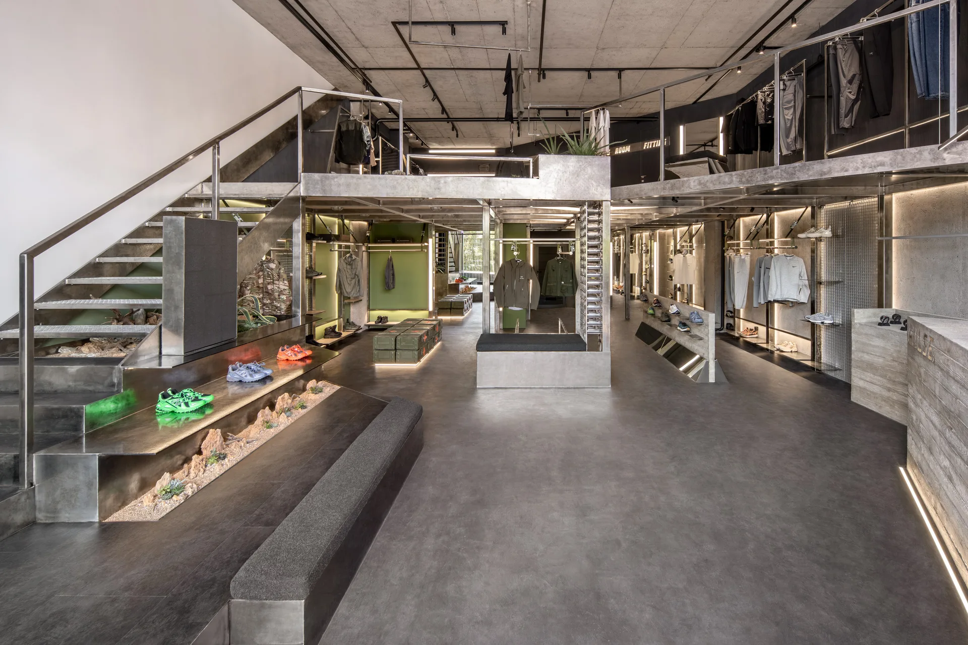 A modern industrial retail space featuring a two-level brushed metal mezzanine, concrete floor and ceiling, and clothing racks with integrated lighting.