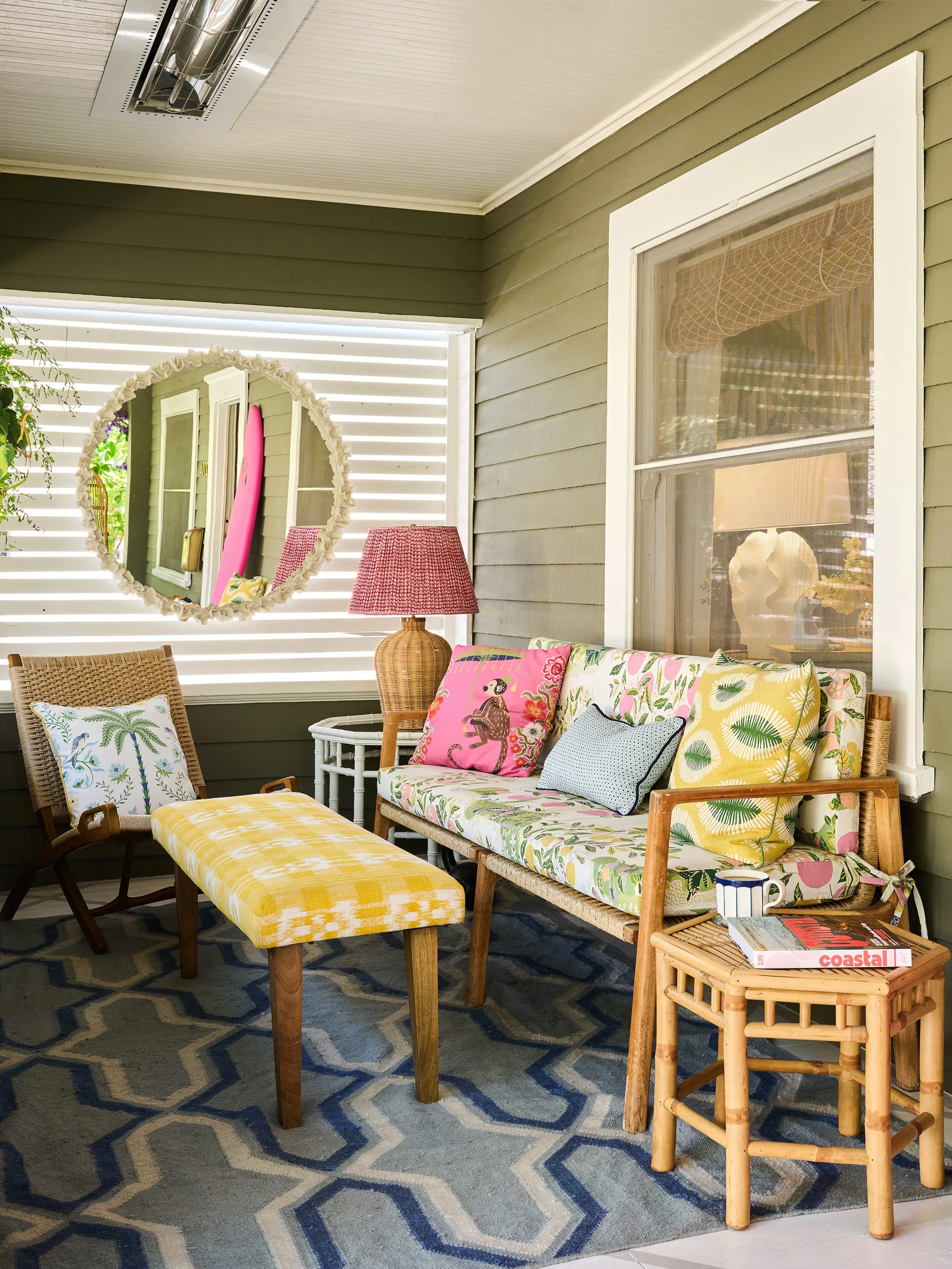 A covered porch featuring olive green siding, a rattan sofa with floral cushions, a yellow plaid bench, and a blue geometric rug.