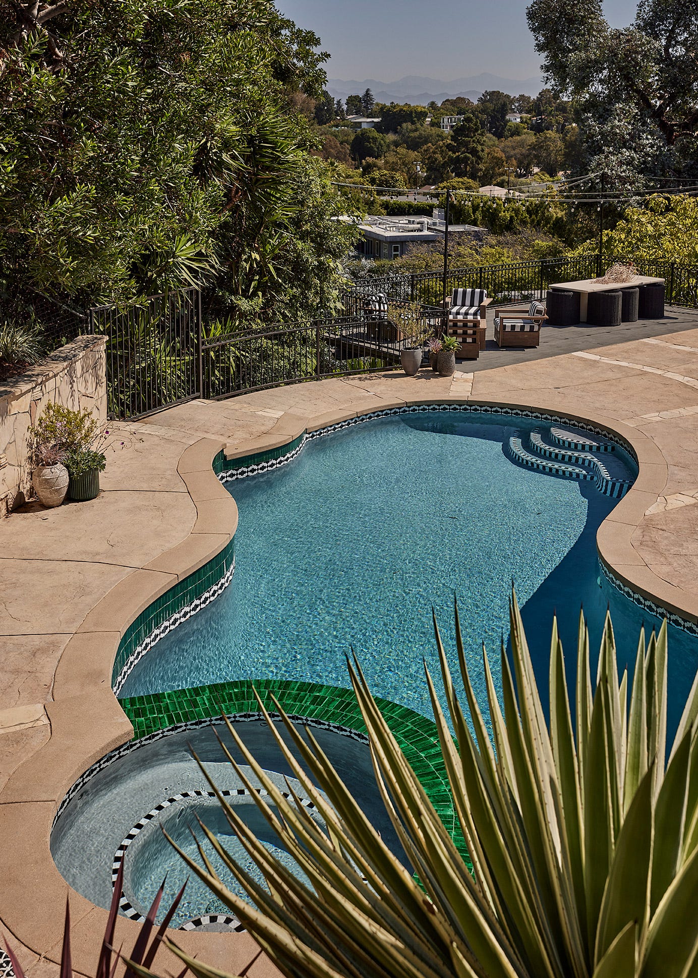 A curved swimming pool with an integrated spa and green tile trim is surrounded by a concrete deck overlooking a hillside landscape.