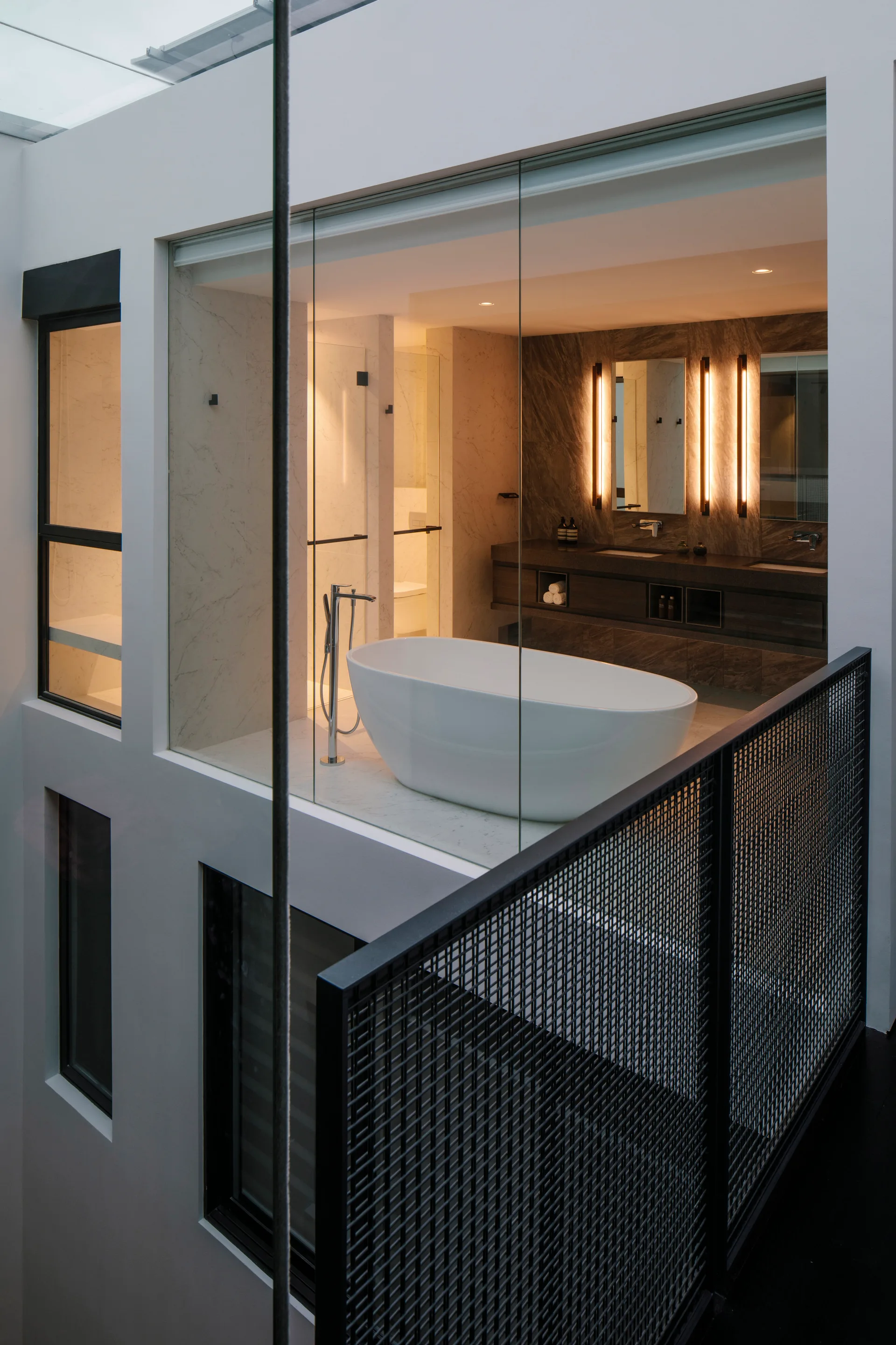 A modern bathroom seen through a glass partition featuring a white freestanding bathtub, dark stone vanity, and vertical light sconces.