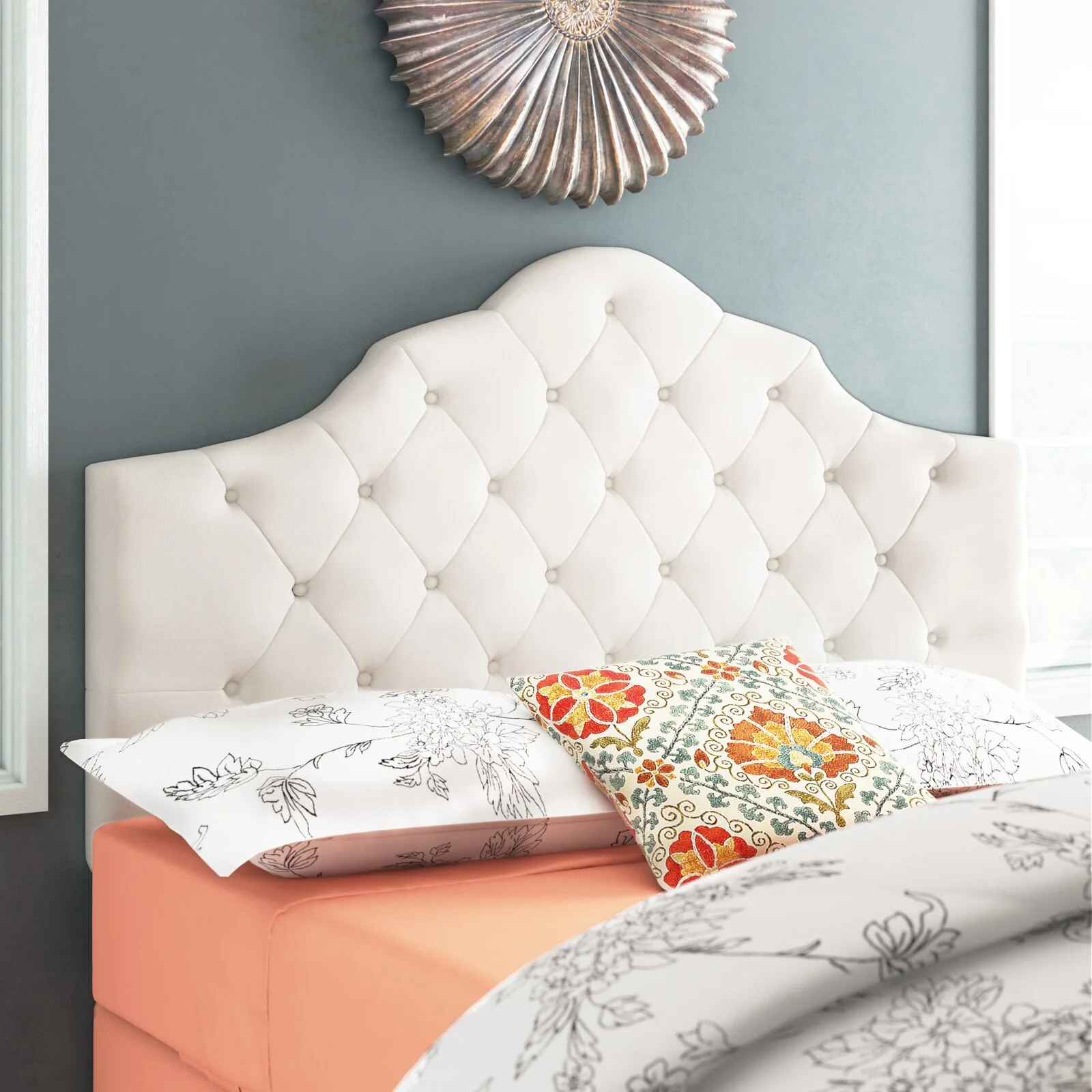 A cream upholstered headboard with button tufting sits against a grey wall next to floral patterned pillows and a metallic wall decoration.