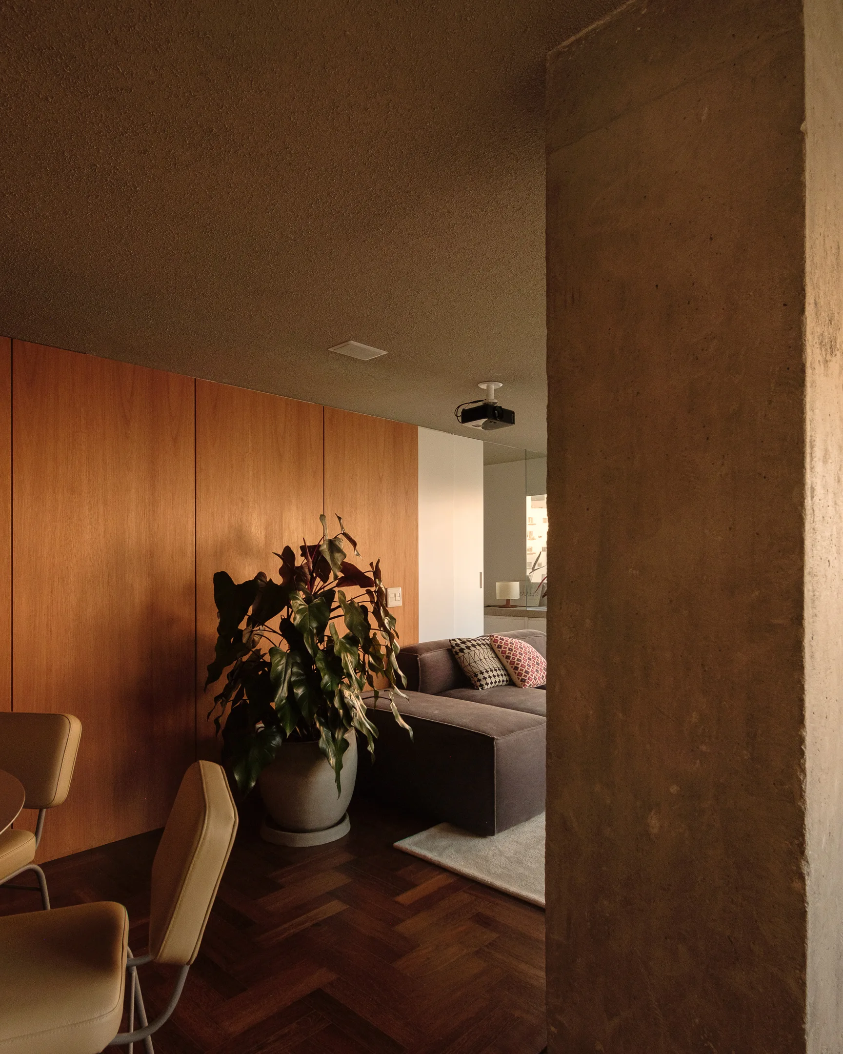An interior residential space features vertical wood wall paneling, an exposed concrete pillar, dark herringbone floors, and a grey sofa.