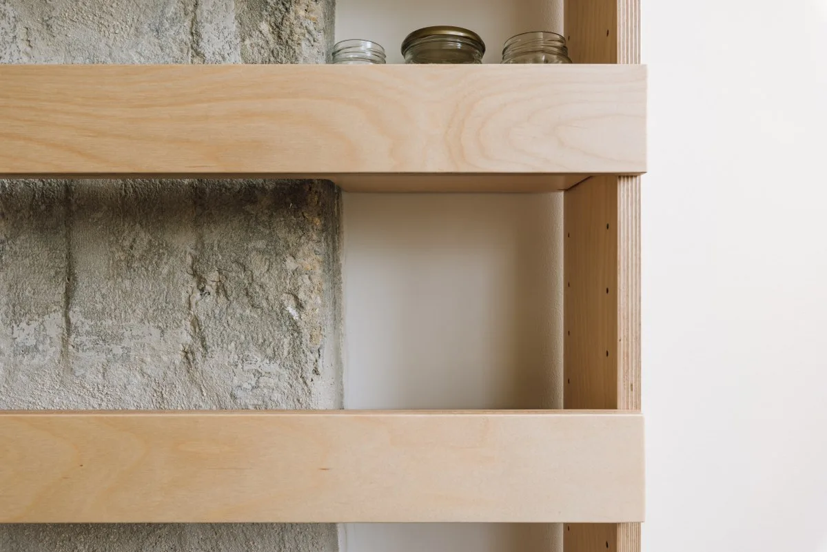 Close-up of a light-colored plywood shelving unit with exposed edges mounted against a textured grey stone wall and a smooth white wall.