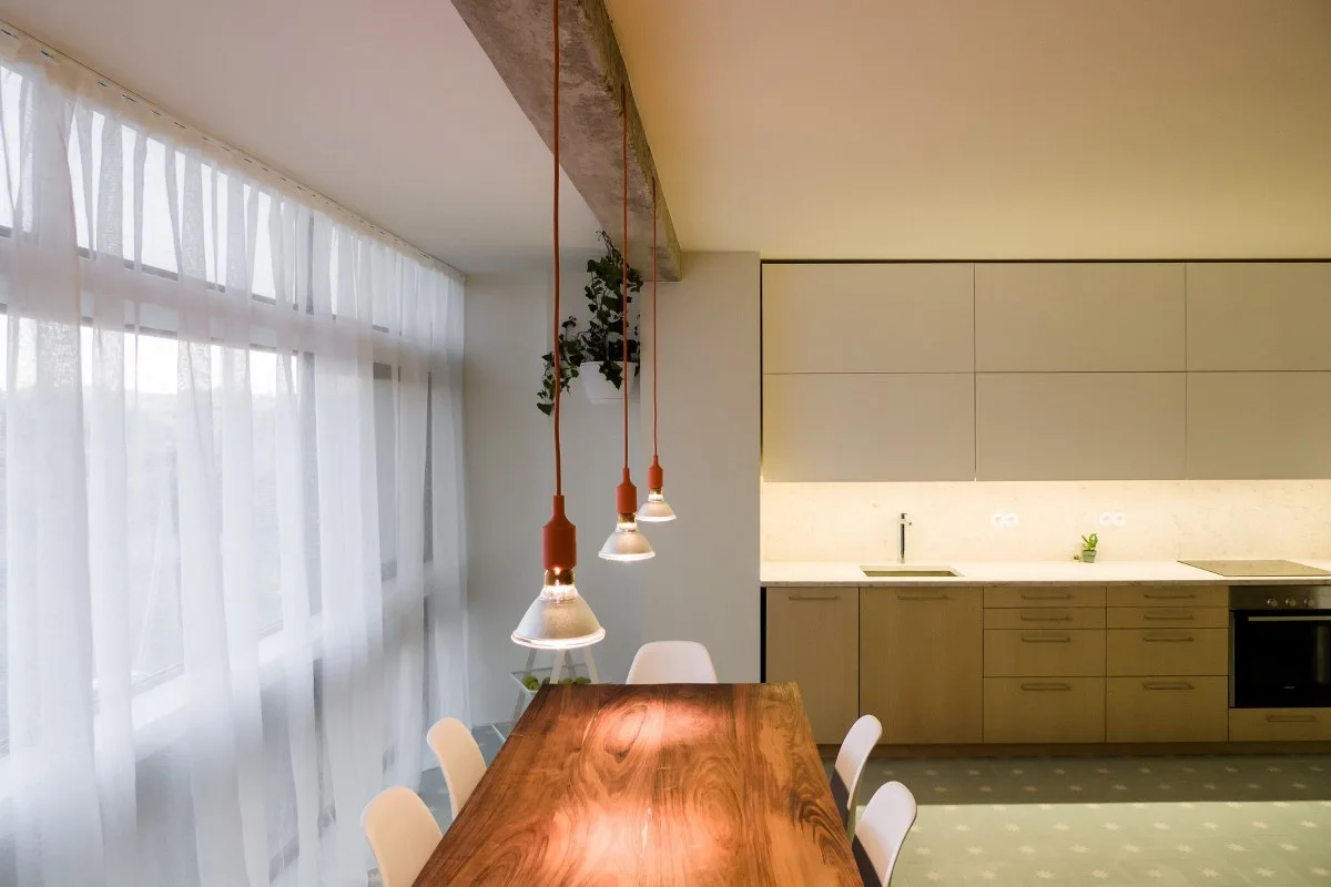 A modern kitchen and dining space with a natural wood slab table, white sheer curtains, and three pendant lights hanging from a concrete beam.