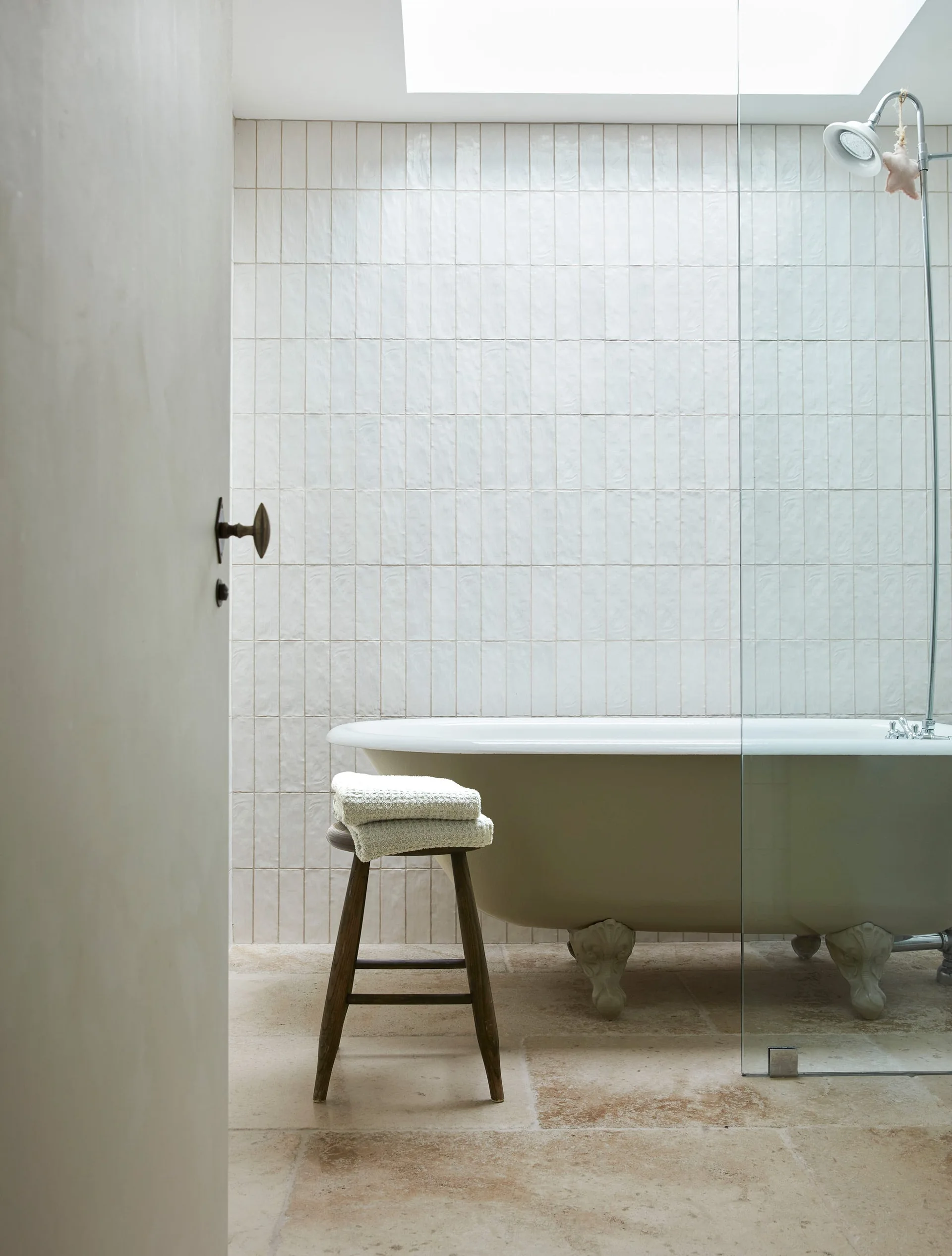 A white clawfoot bathtub stands on a stone floor in front of a vertically stacked tile wall under a bright skylight.