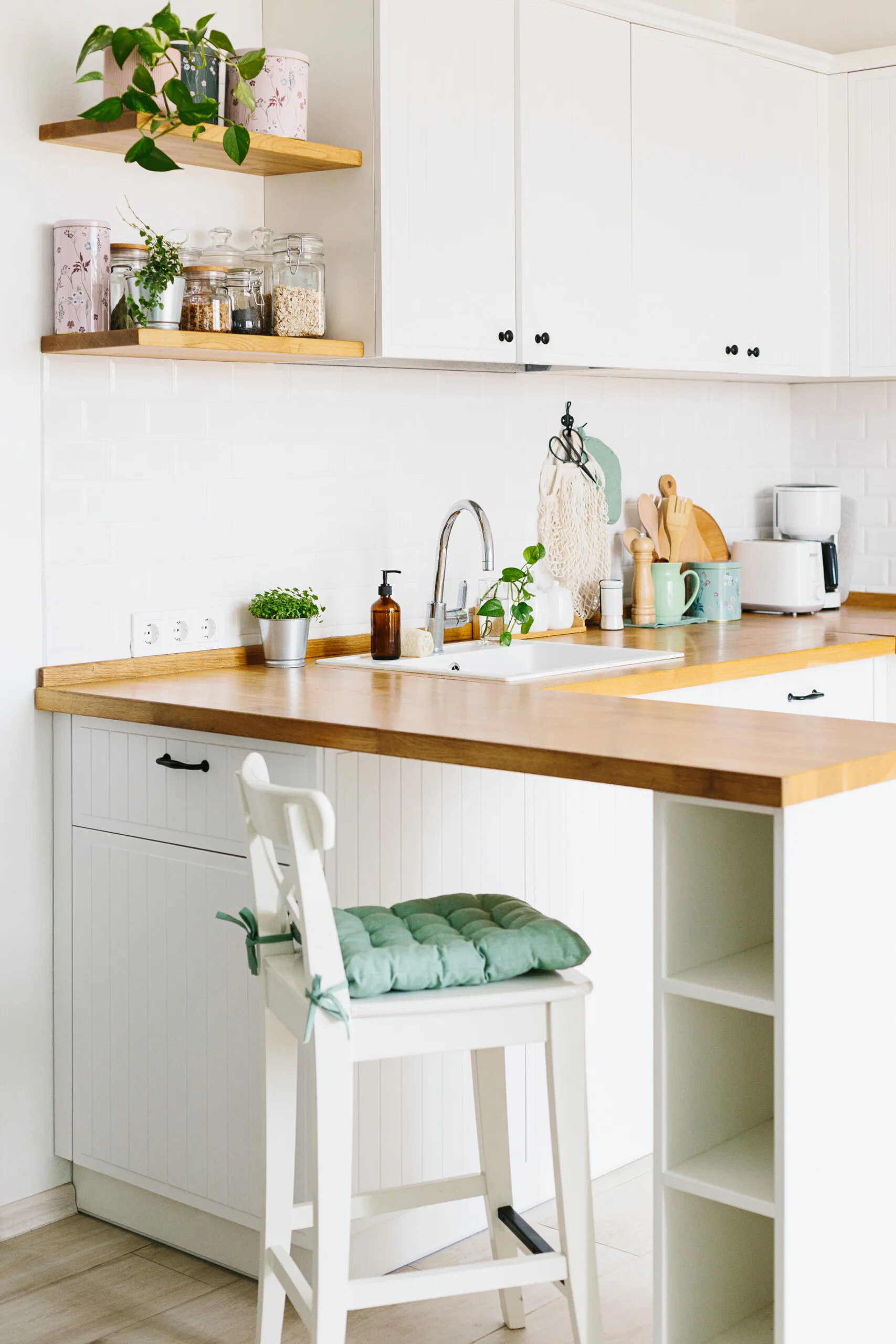 White kitchen cabinetry features a wooden countertop, subway tile backsplash, floating wood shelves, and a white bar stool with a green cushion.