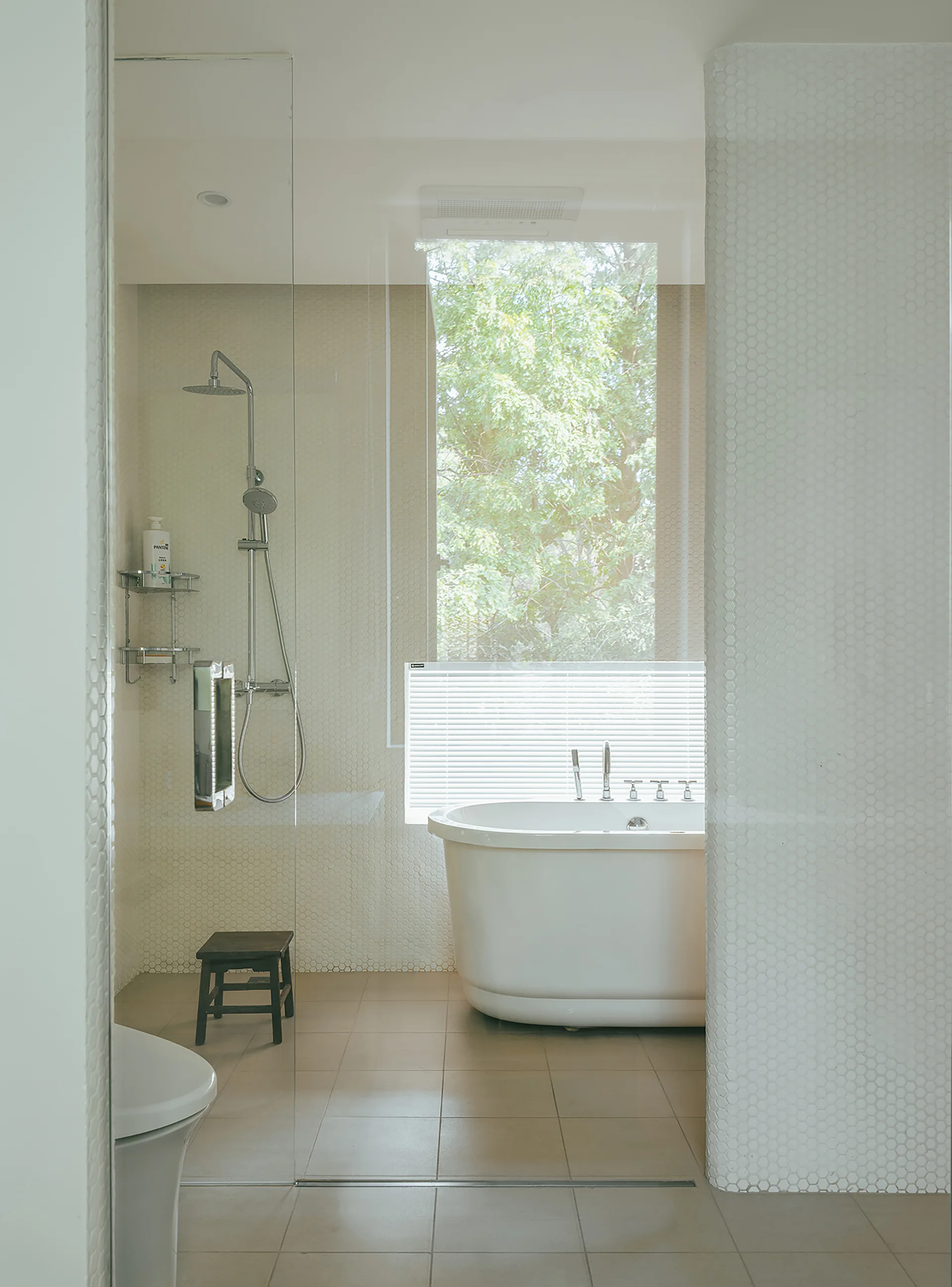 A minimalist bathroom with a white soaking tub by a window, hexagonal mosaic tile walls, and a glass partition separating the shower area.