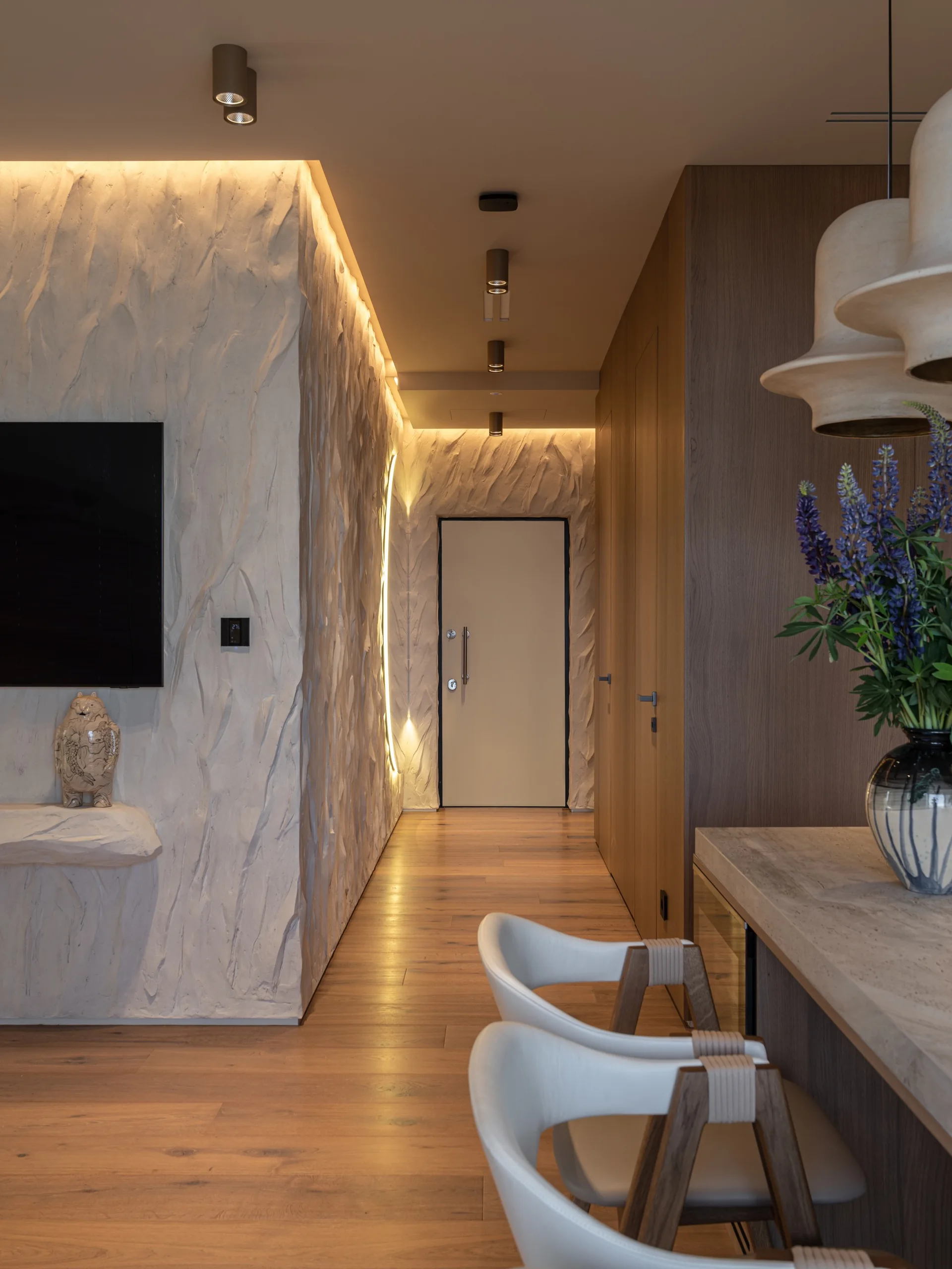 A modern hallway features a heavily textured plaster wall on one side, seamless wood-grain cabinets on the other, and warm recessed ceiling lights.