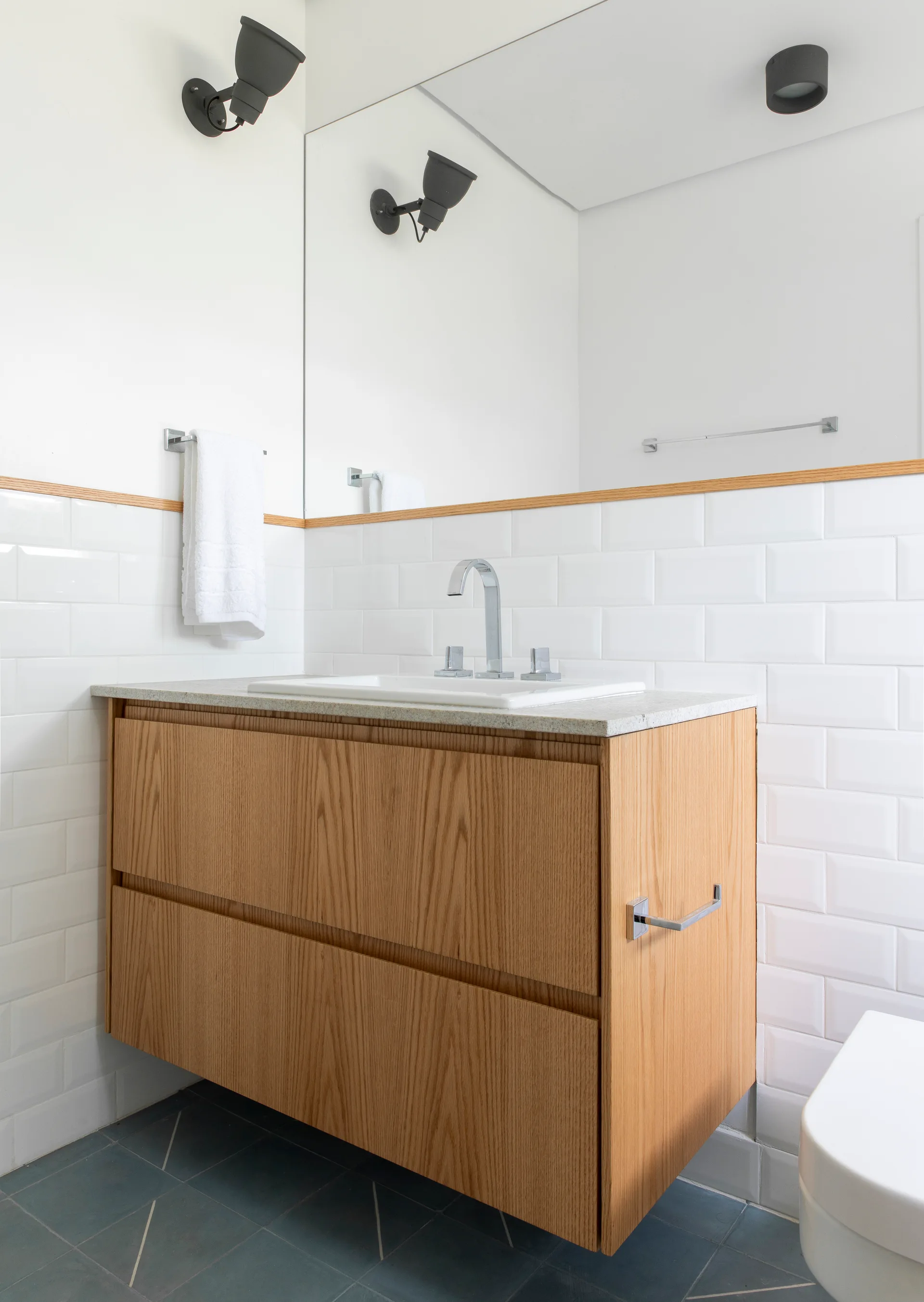 A floating oak bathroom vanity with a white sink is mounted against a white subway tile wall beneath a large mirror and black sconces.