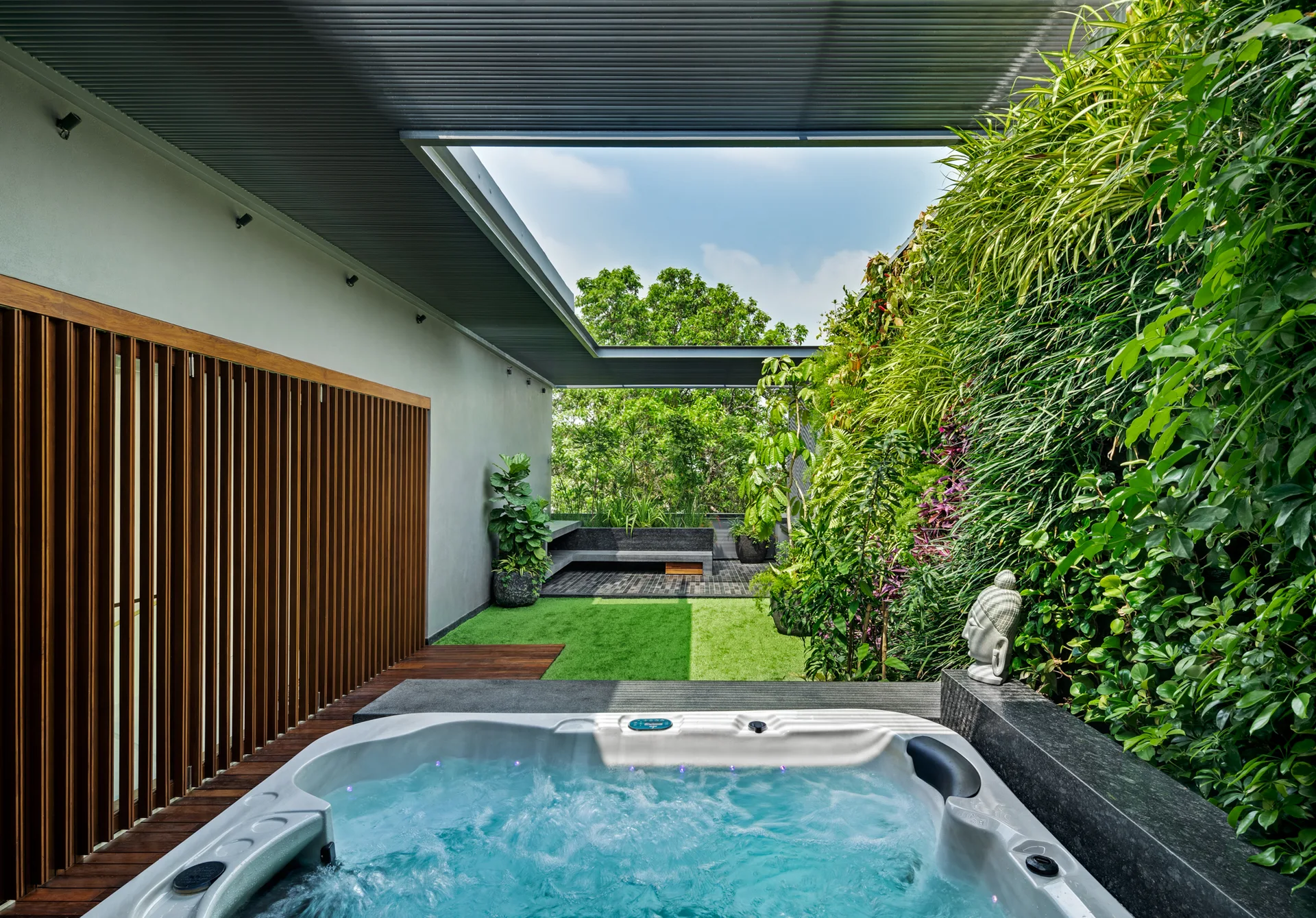 A modern outdoor spa patio featuring a bubbling hot tub next to a lush vertical garden wall, wood slats, and a slatted ceiling overhang.