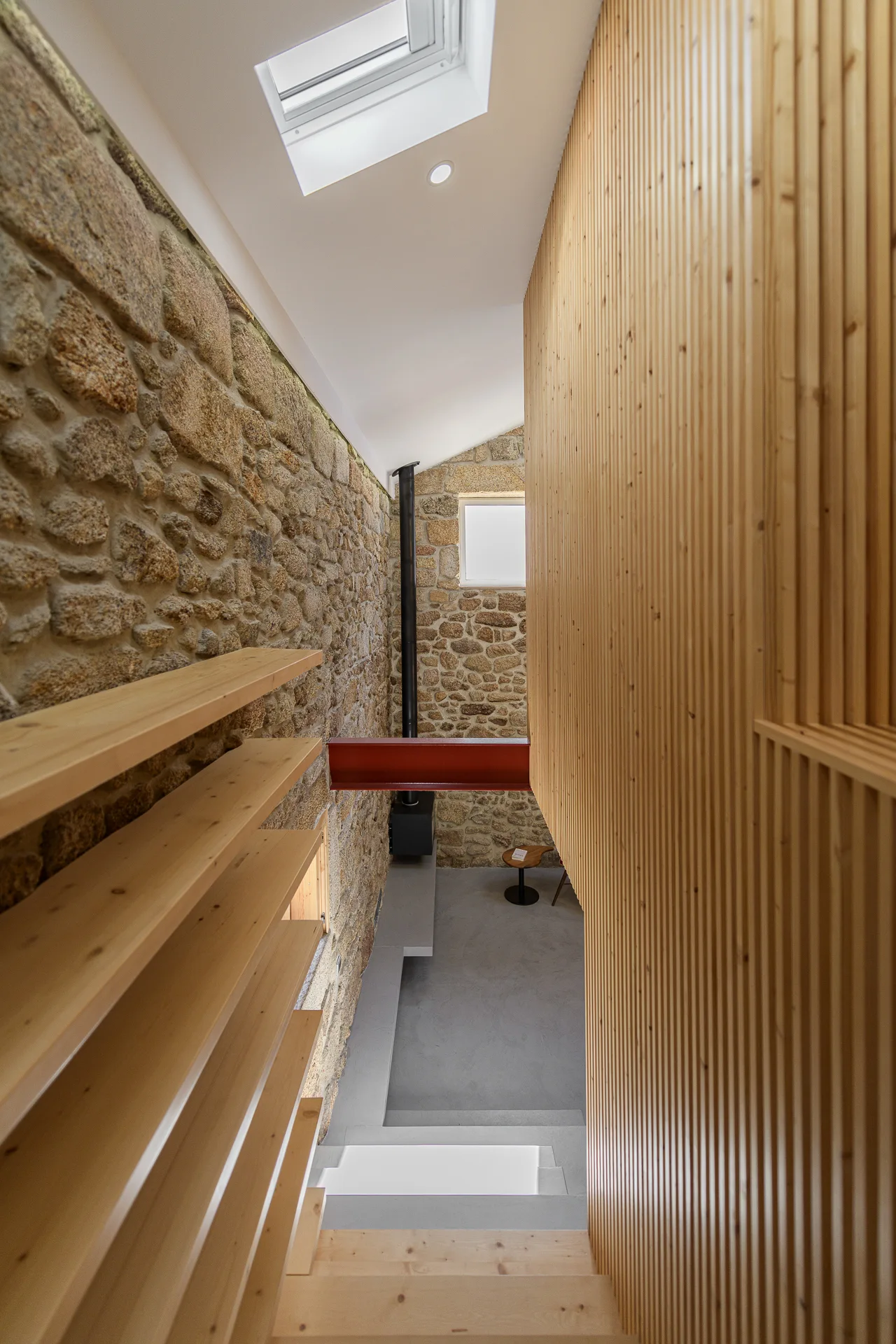 A wooden staircase descends between a textured natural stone wall with open shelves and a modern vertical wood slatted wall under a skylight.
